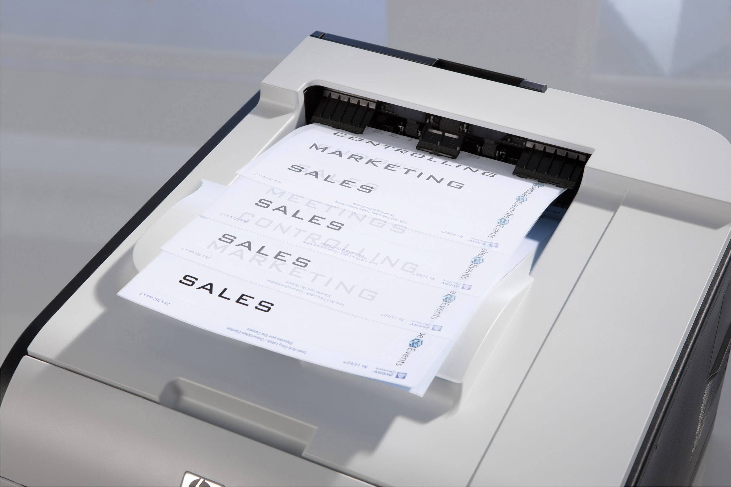 Ein Drucker gibt mehrere Seiten mit dem Wort 'SALES' aus, die auf einem Ausgabefach gestapelt sind. Das Bild zeigt den Druckprozess.