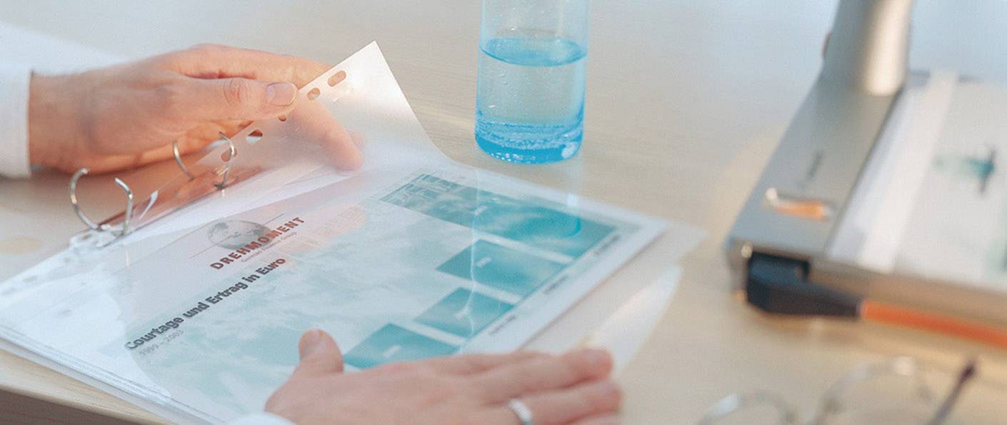 Eine Person blättert durch ein Dokument in einer Klarsichtfolie auf einem Schreibtisch, neben einem Laptop und einem Wasserglas.