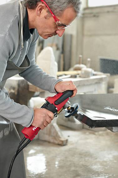 Ein Mann schleift mit einem Winkelschleifer die Kante einer Steinplatte in einer Werkstatt. Trägt Schutzbrille und Arbeitskleidung.