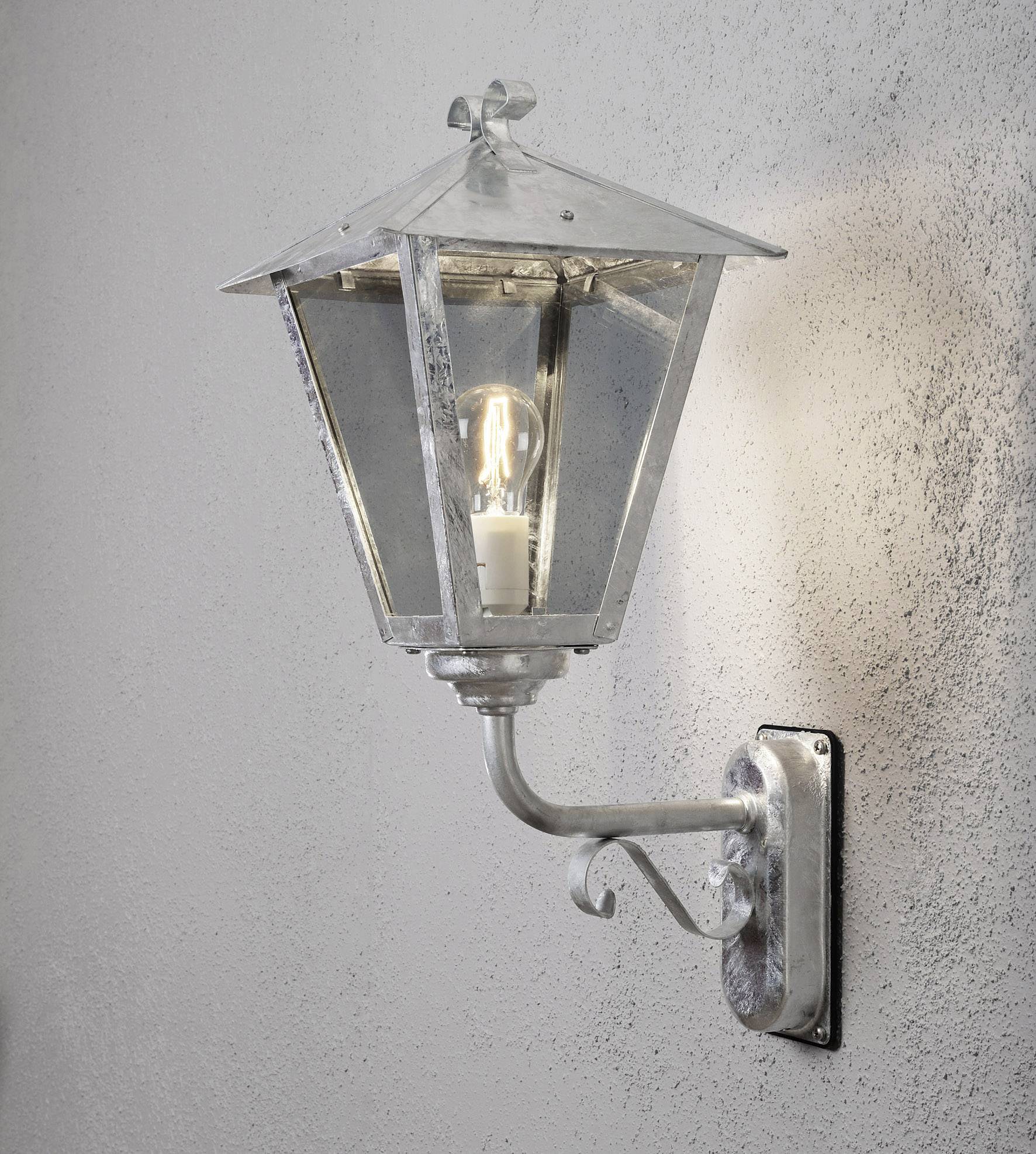 Wandleuchte aus Metall mit Glas, an einer strukturierten Wand montiert. Sie hat eine klassische Laternenform und strahlt warmes Licht aus.