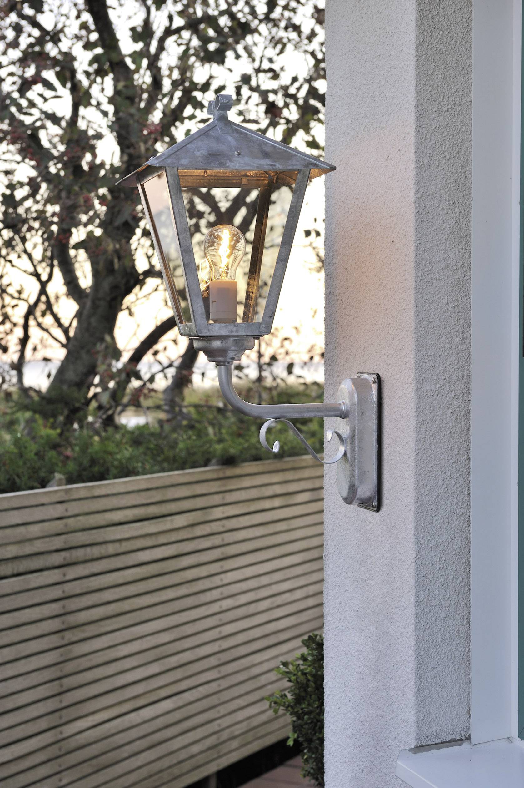 Wandleuchte im traditionellen Stil leuchtet bei Dämmerung. Sie ist an einer Hauswand montiert, mit einem Baum im Hintergrund.