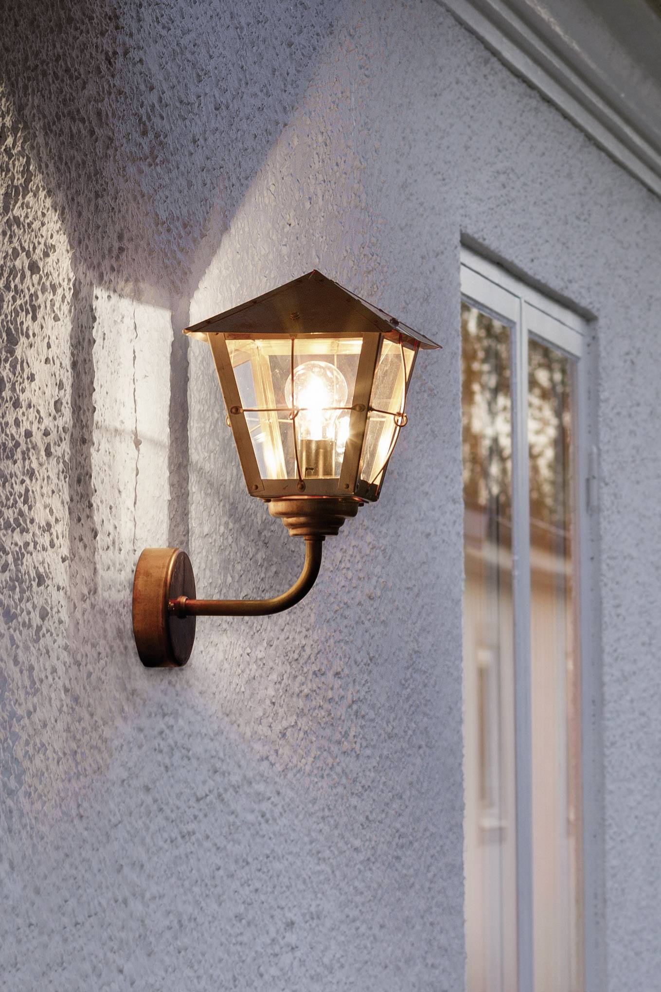 Eine eingeschaltete Wandlampe mit Glühbirne leuchtet an einer Außenwand neben einem Fenster.