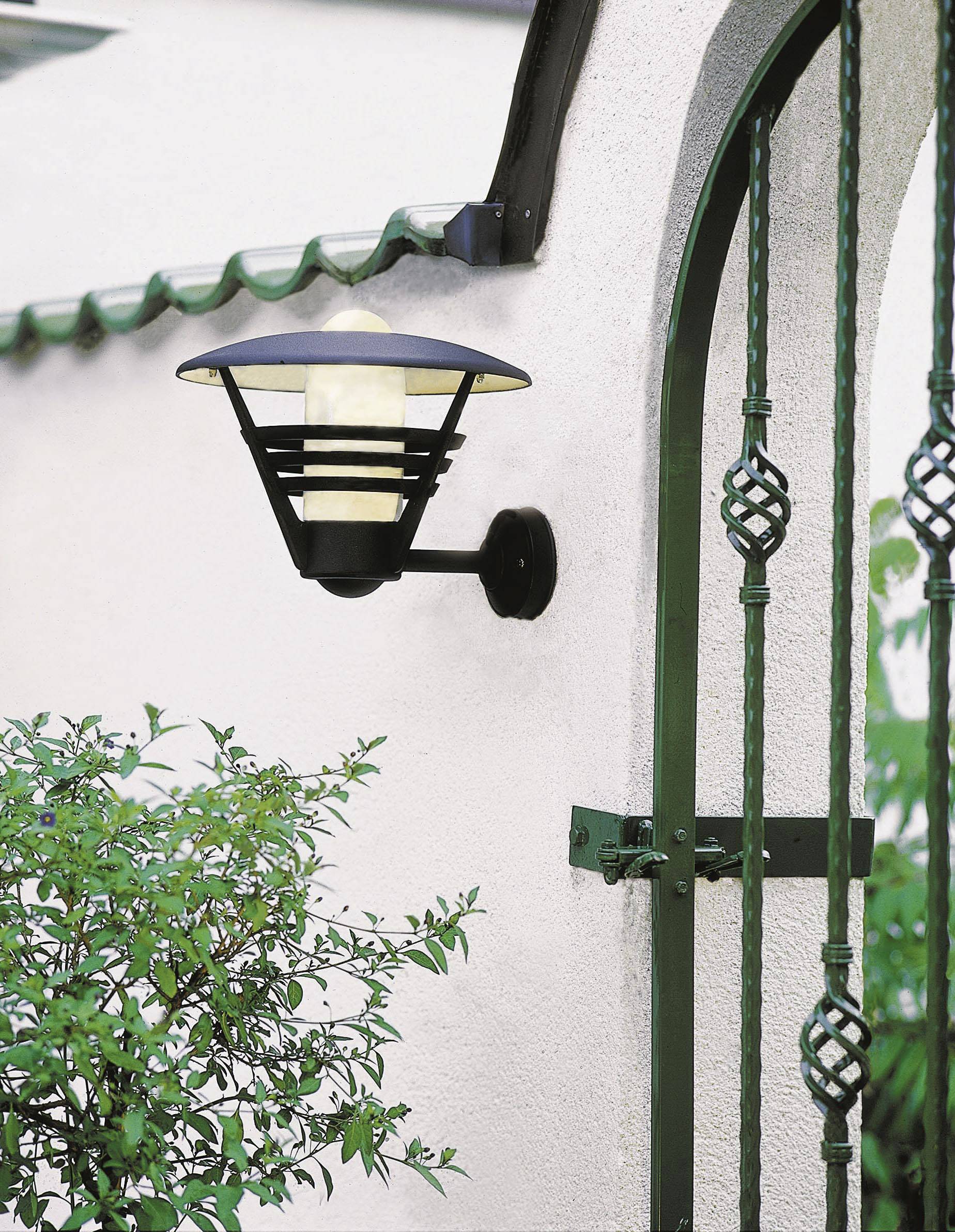 Wandlampe an weißer Wand, neben einem grünen Zaun, gibt sanftes Licht ab.