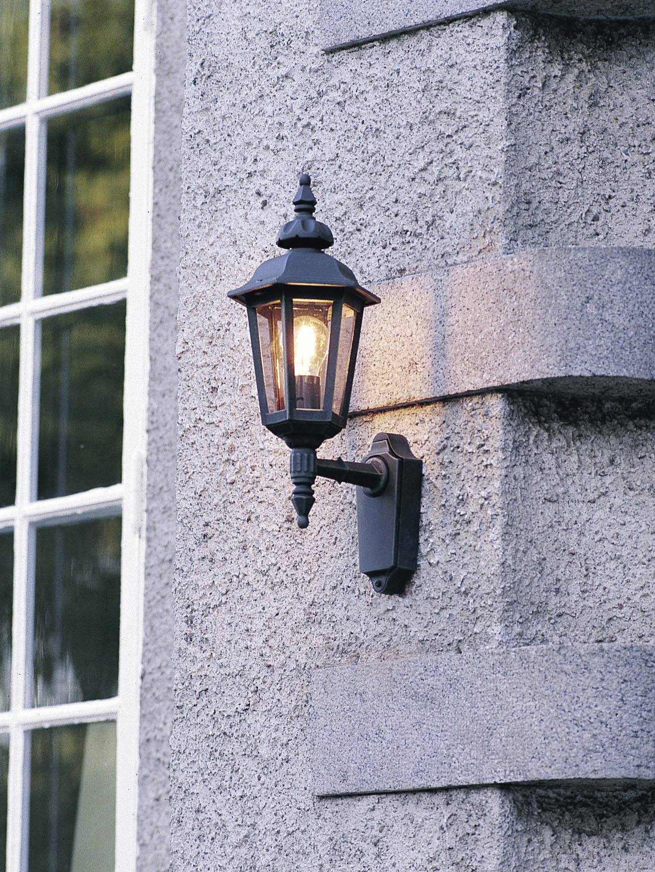 Außenwandlaterne aus schwarzem Metall mit beleuchteter Glühlampe an einer strukturierten Steinwand neben einem Fenster.