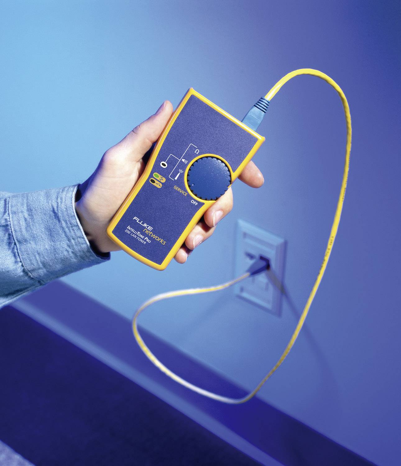 Eine Hand hält ein gelbes elektrisches Messgerät mit einem Kabel, das an eine Wandsteckdose angeschlossen ist.