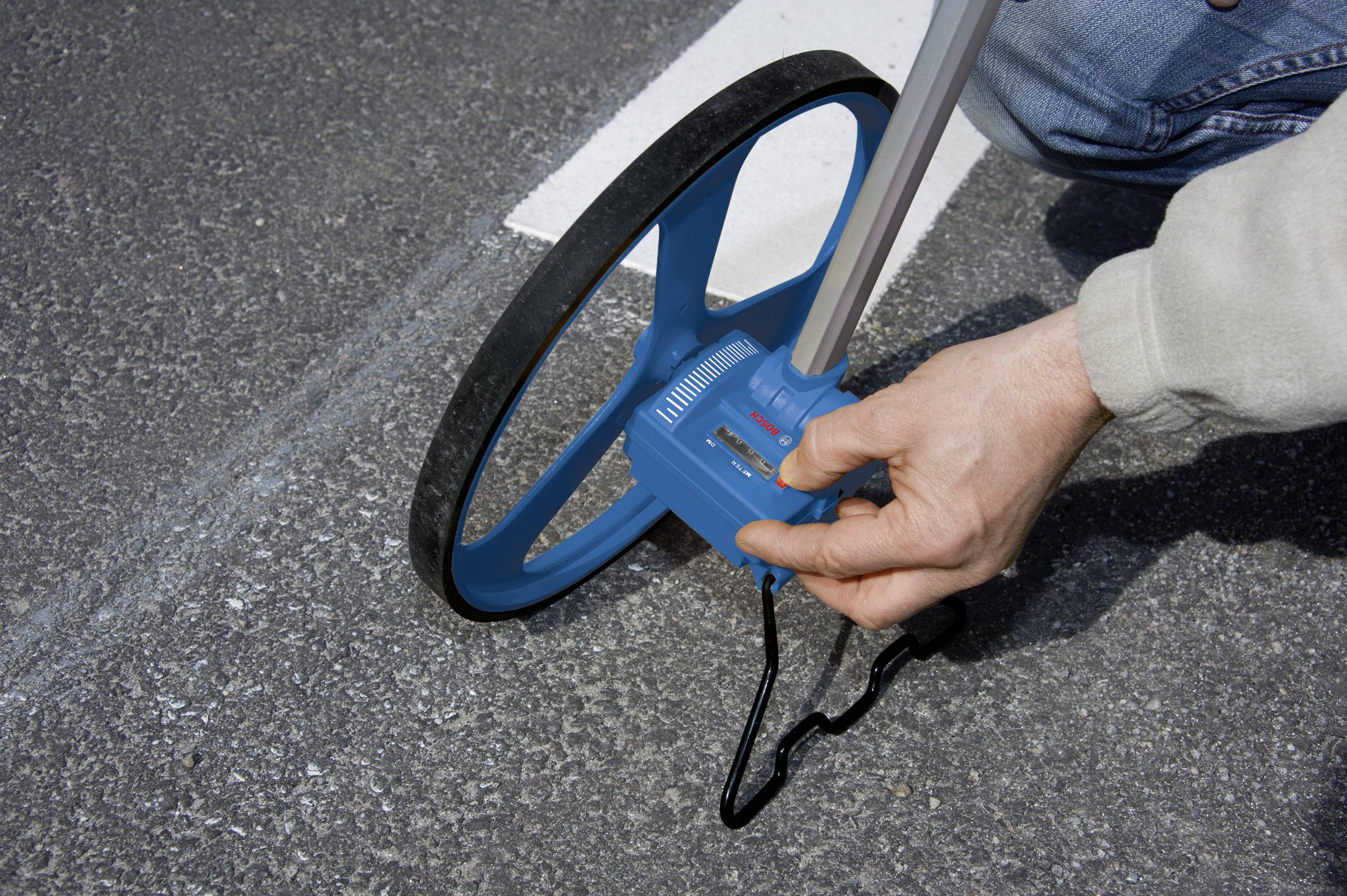 Eine Person misst mit einem Rolltacho eine Asphaltlänge entlang einer weißen Linie.