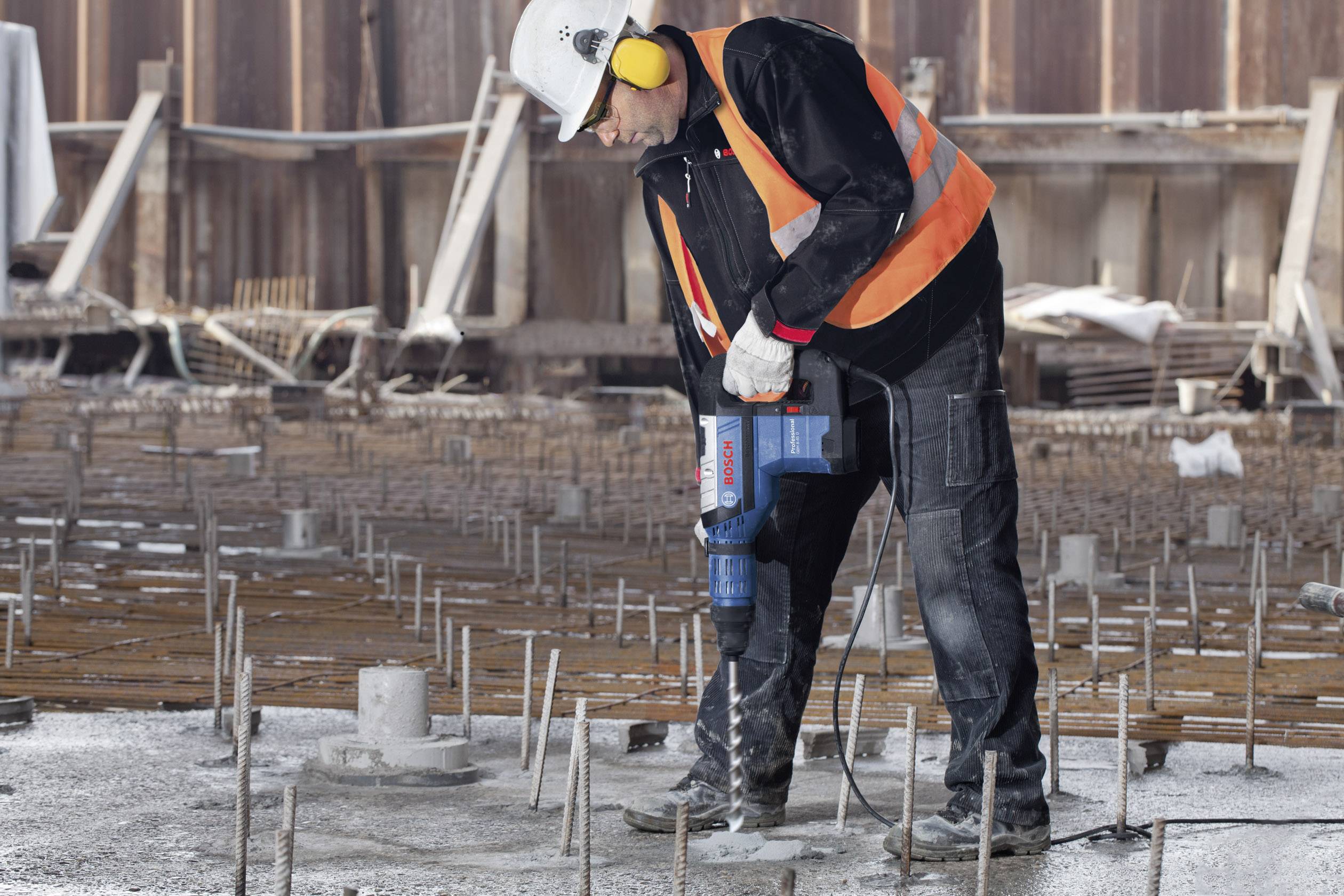 Ein Bauarbeiter mit Schutzhelm und Gehörschutz bohrt mit einer elektrischen Bohrmaschine in eine Betonplatte auf einer Baustelle.