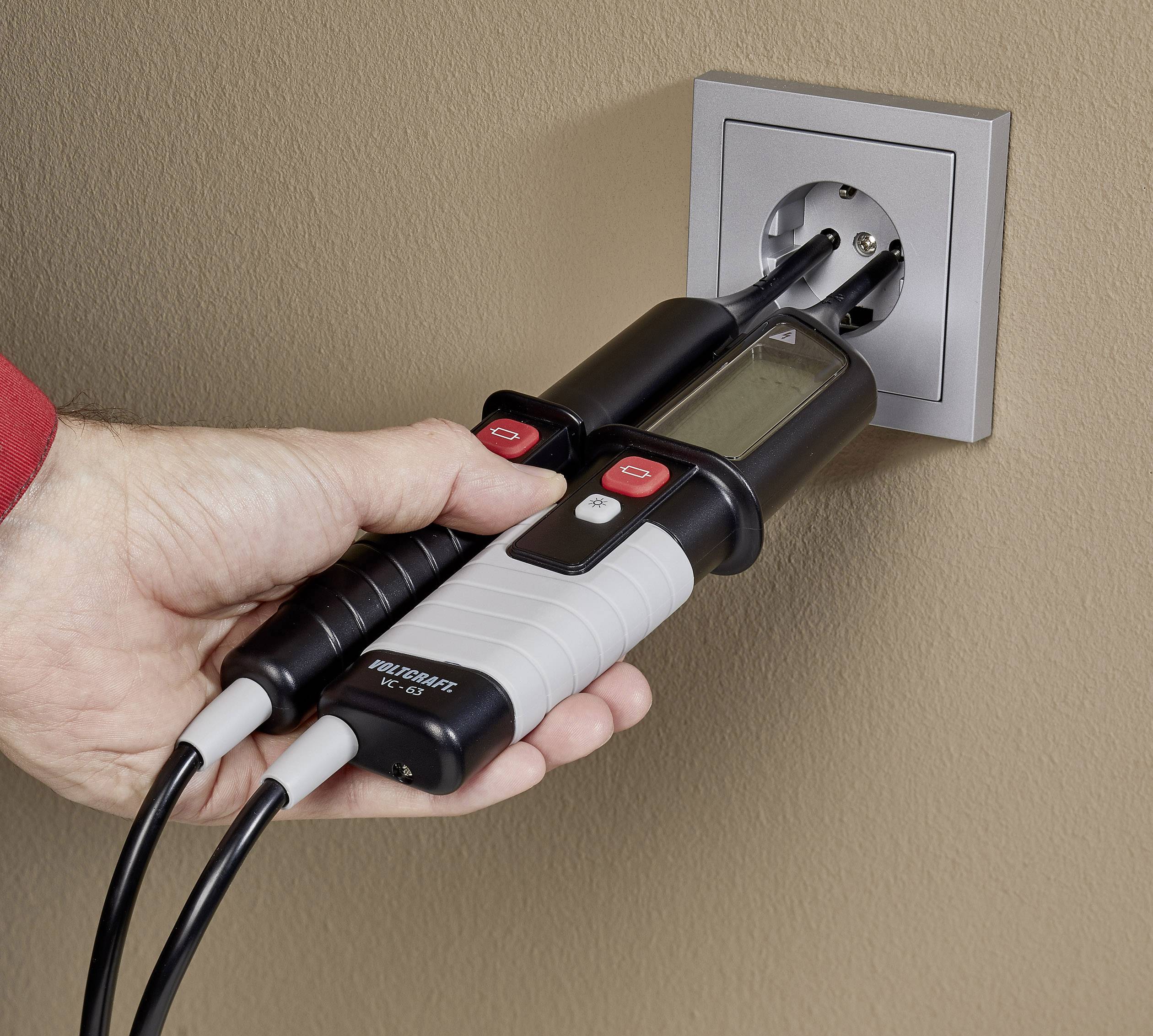 Eine Hand testet mit einem Spannungsprüfer eine Steckdose. Der Prüfer zeigt eine digitale Anzeige. Die Wand ist beige.