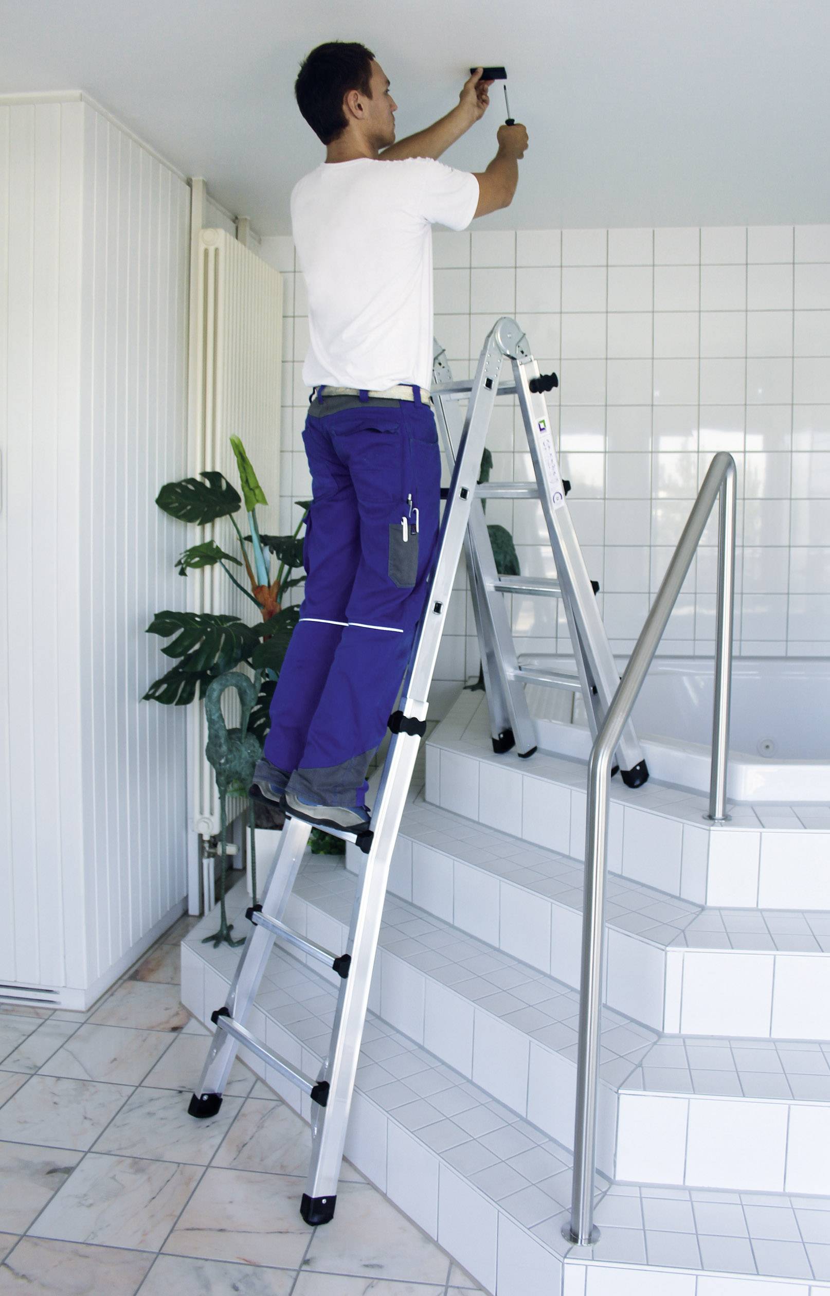 Eine Person in Arbeitskleidung steht auf einer Leiter und arbeitet an der Decke in einem gefliesten Raum mit Pflanzen.