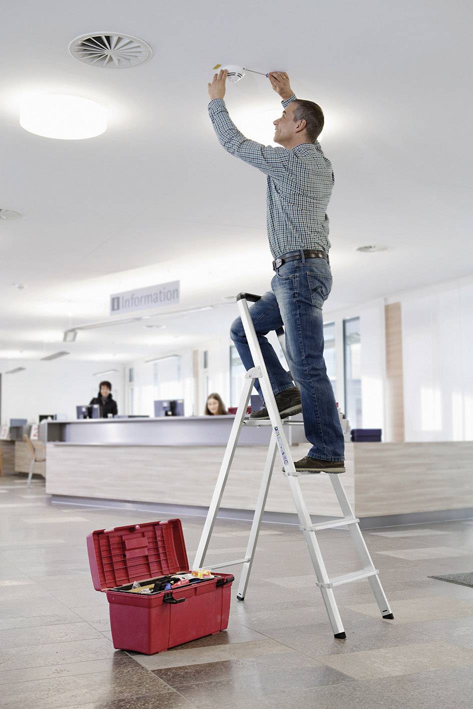 Ein Mann steht auf einer Leiter und repariert eine Deckenlampe in einem Büro. Daneben steht ein roter Werkzeugkasten. Im Hintergrund ist ein Informationsschalter.
