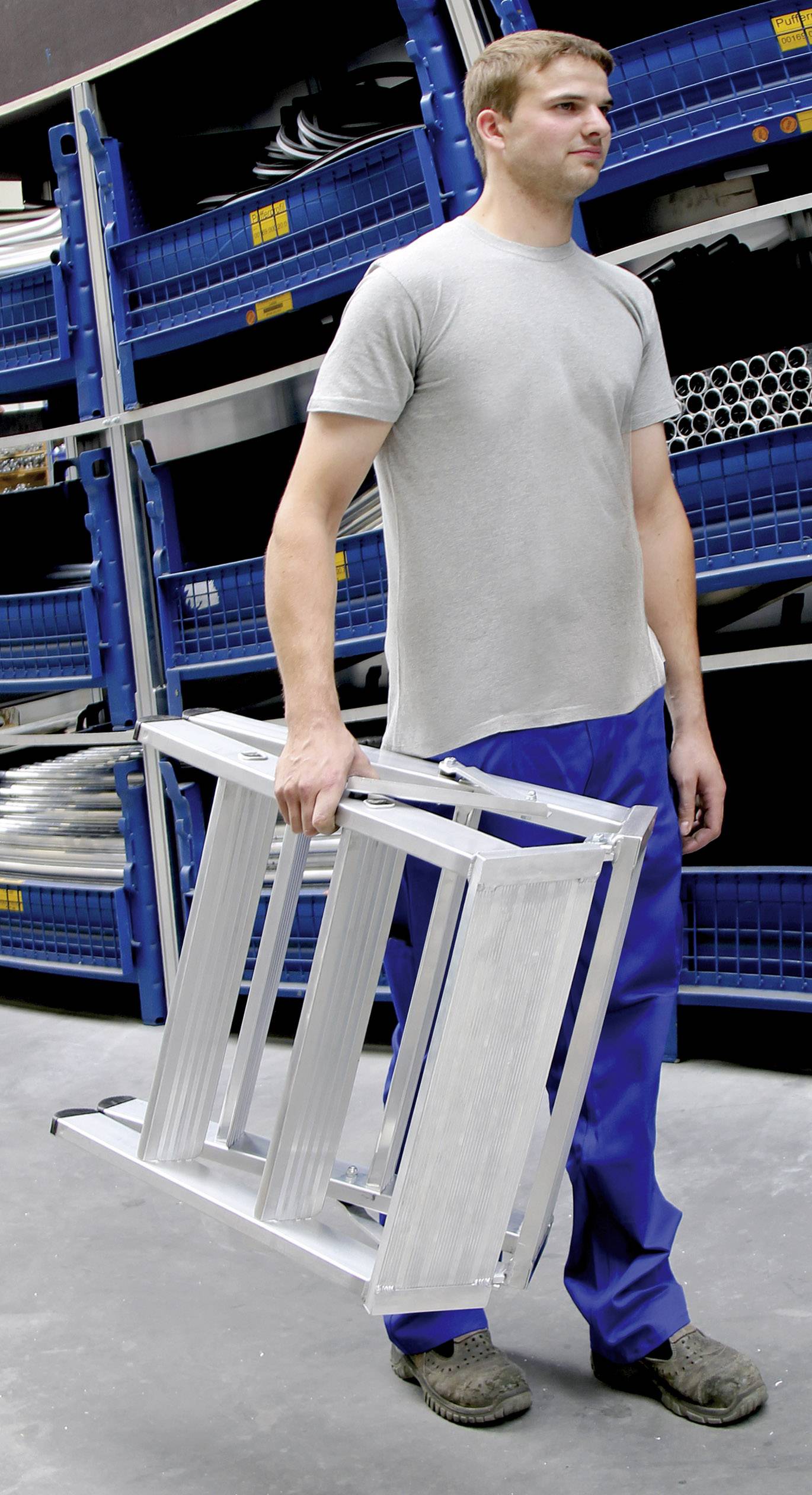 Ein Mann in Arbeitskleidung trägt eine Aluminiumleiter in einem Lager mit blauen Regalen im Hintergrund.