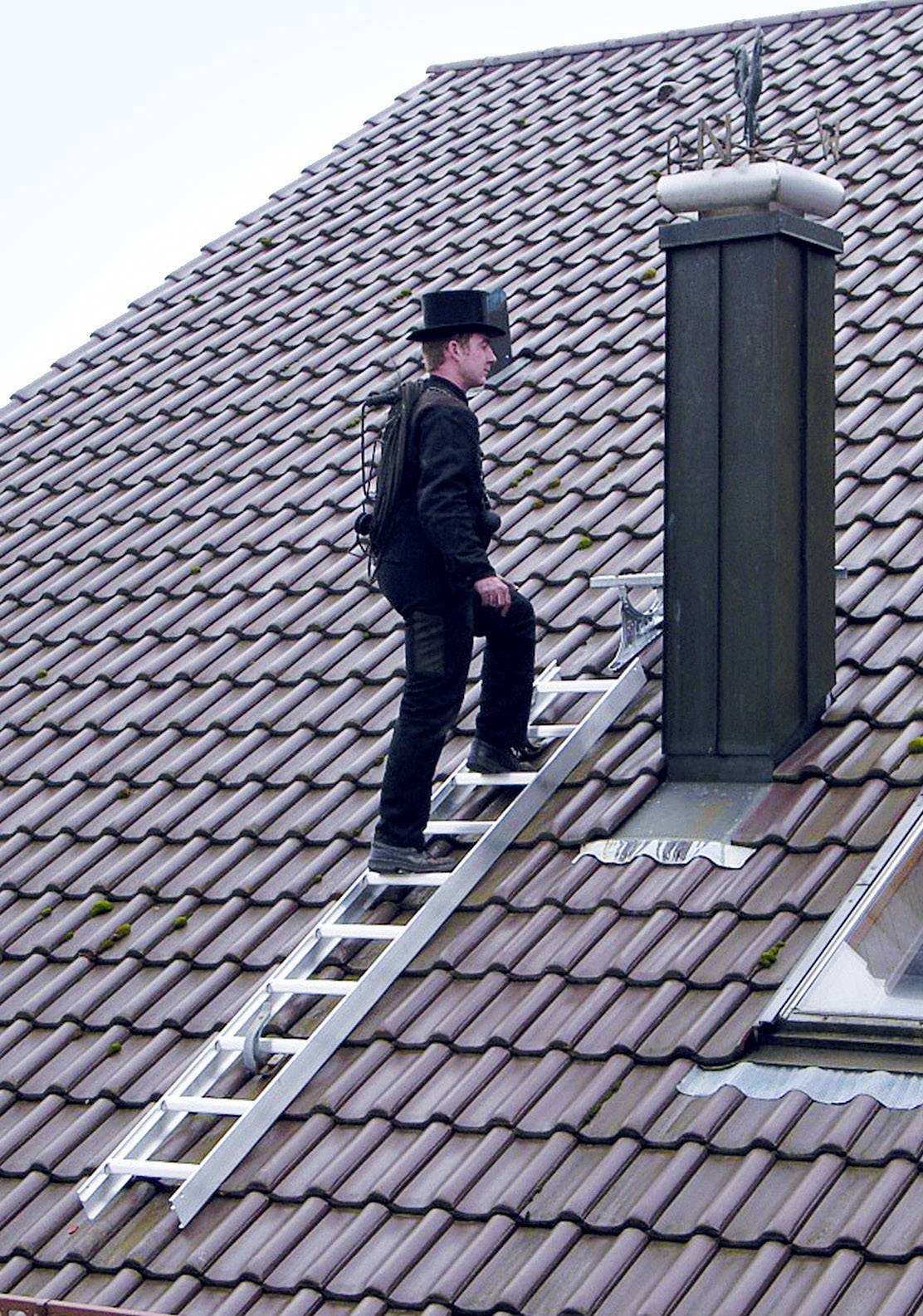 Ein Schornsteinfeger in traditioneller Kleidung steigt eine Leiter hinauf, um einen hohen Schornstein auf einem Ziegeldach zu erreichen.
