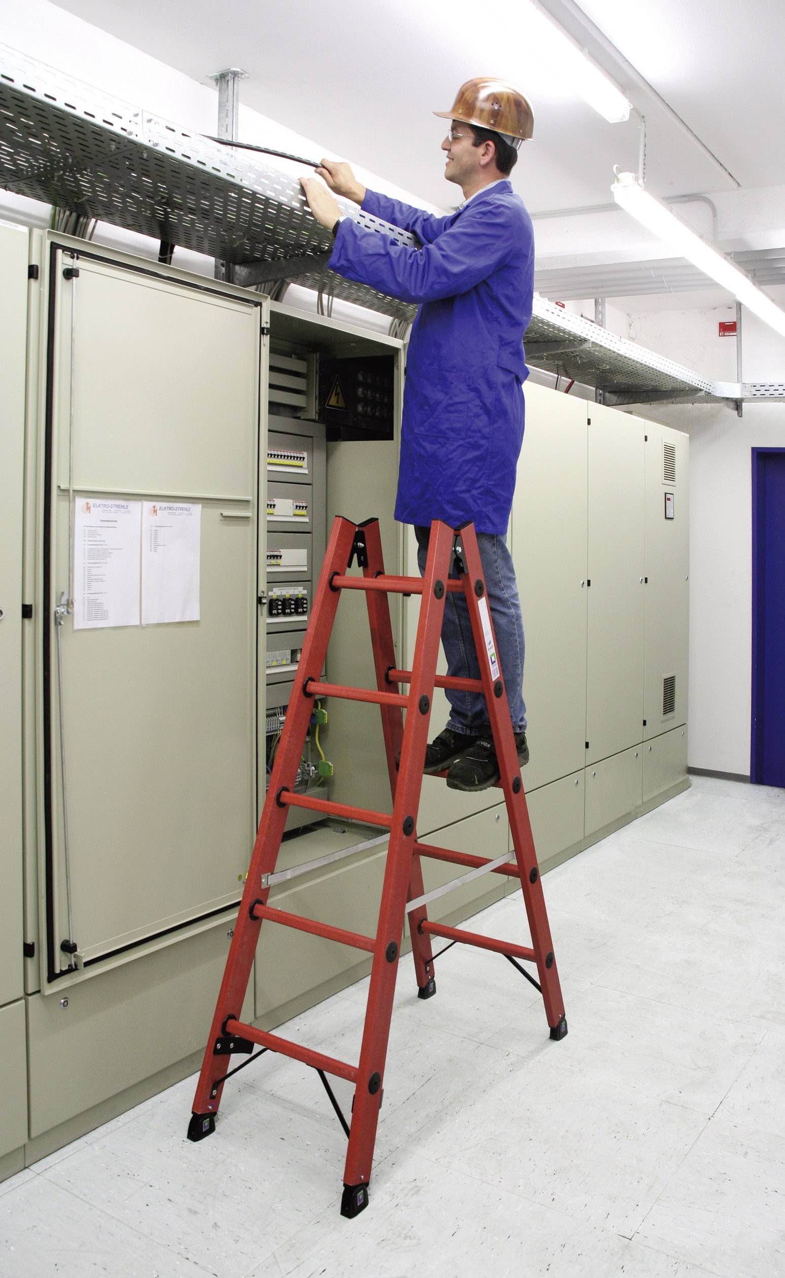 Eine Person in Arbeitskleidung steht auf einer Leiter und arbeitet an einem Stromkabel über großen Schaltschränken in einem Technikraum.