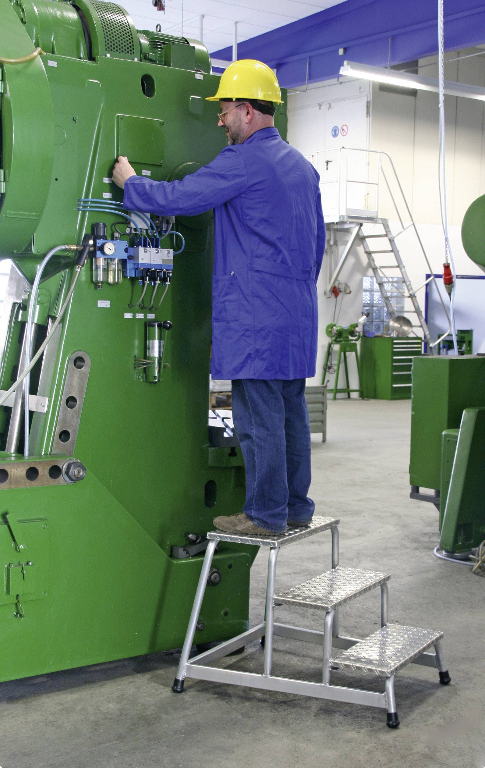 Ein Mann in blauer Arbeitskleidung und gelbem Schutzhelm steht auf einer Leiter und bedient eine große grüne Industriemaschine.