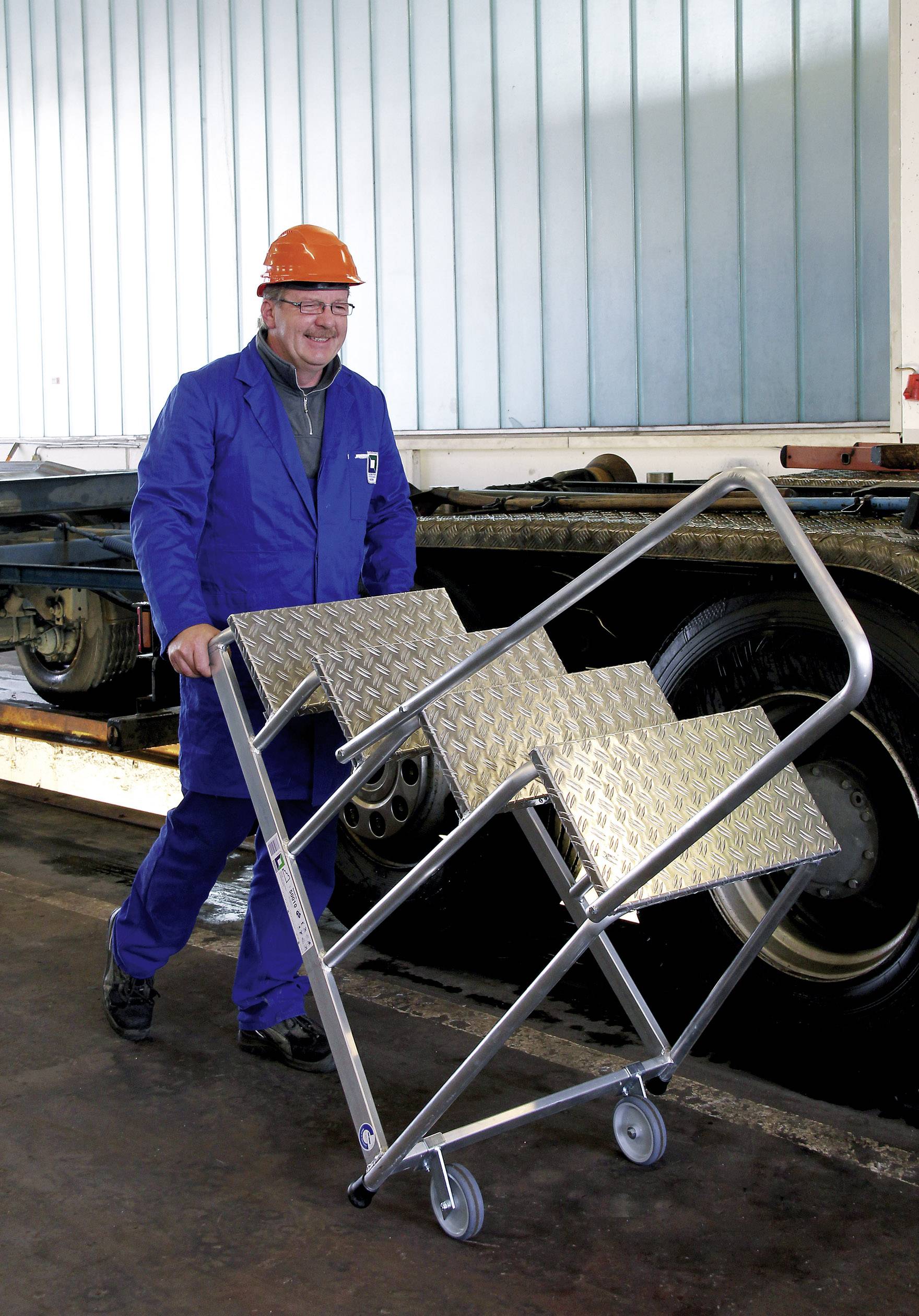 Ein Mann in Arbeitskleidung und Schutzhelm schiebt eine mobile Treppe auf Rädern in einer industriellen Umgebung.