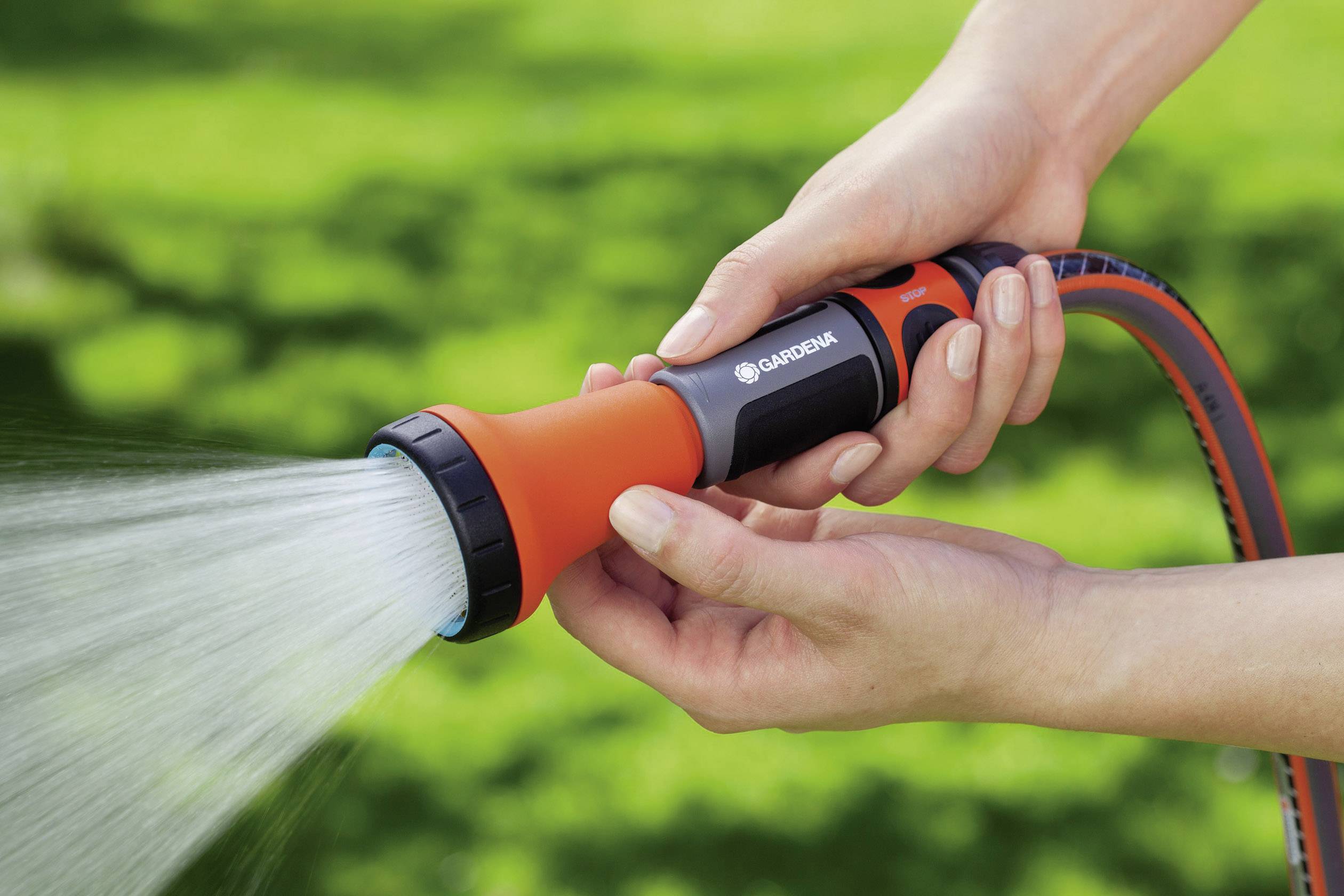 Jemand hält einen Gartenschlauch mit Sprühdüse und bewässert einen Rasen. Der Schlauch ist orange mit schwarzen Details.
