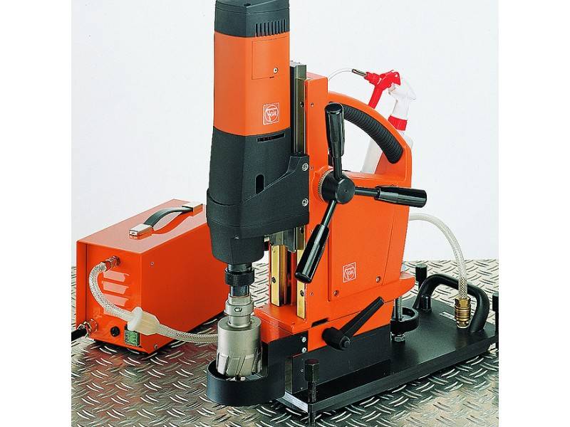 Eine orange-graue magnetische Bohrmaschine auf einer Metallplatte. Sie hat ein Steuergerät und ist mit Kabeln und Schläuchen verbunden.