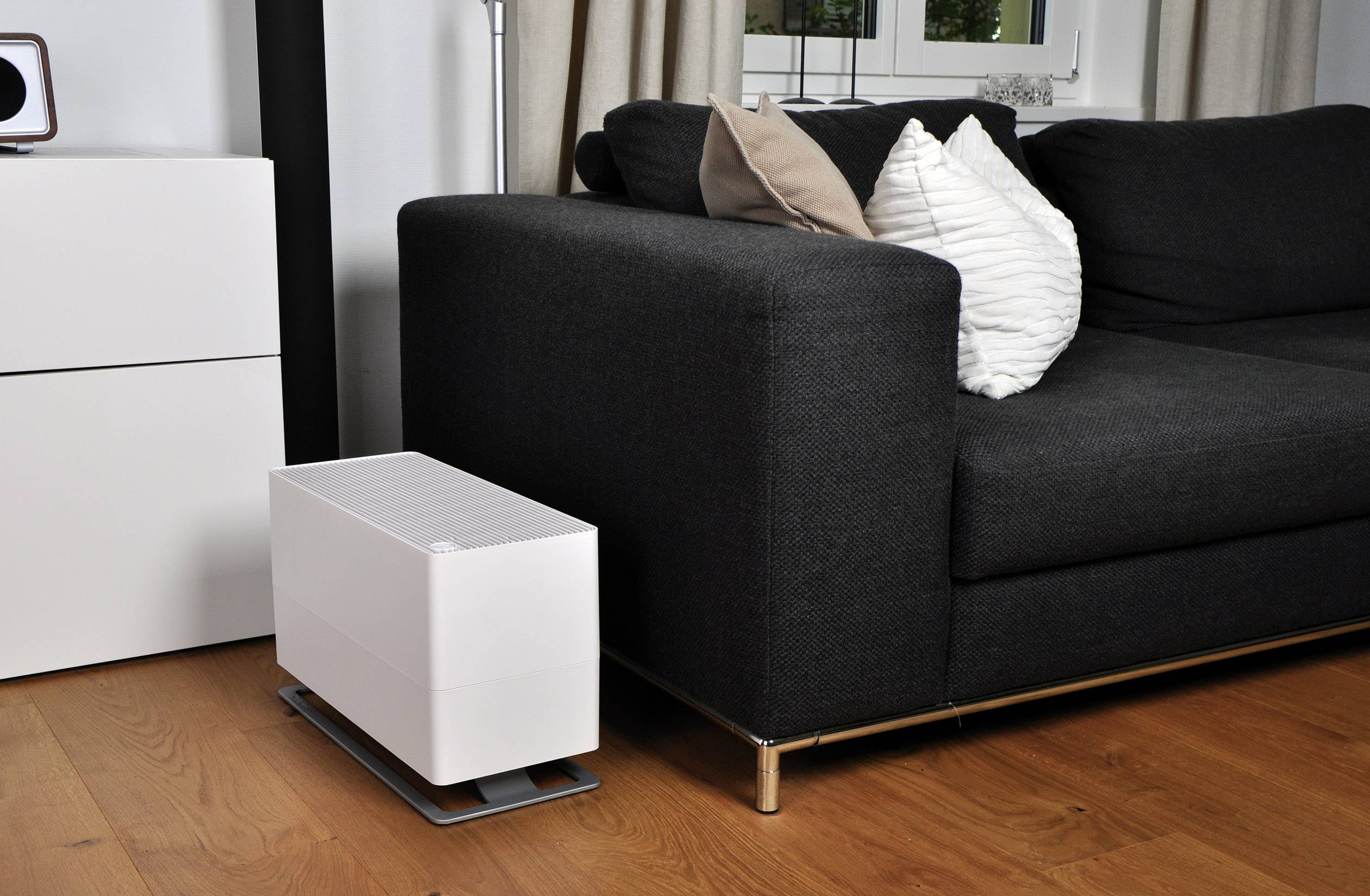 Ein weißer Luftbefeuchter steht auf einem Holzboden neben einem schwarzen Sofa mit weißen und braunen Kissen in einem Wohnzimmer.