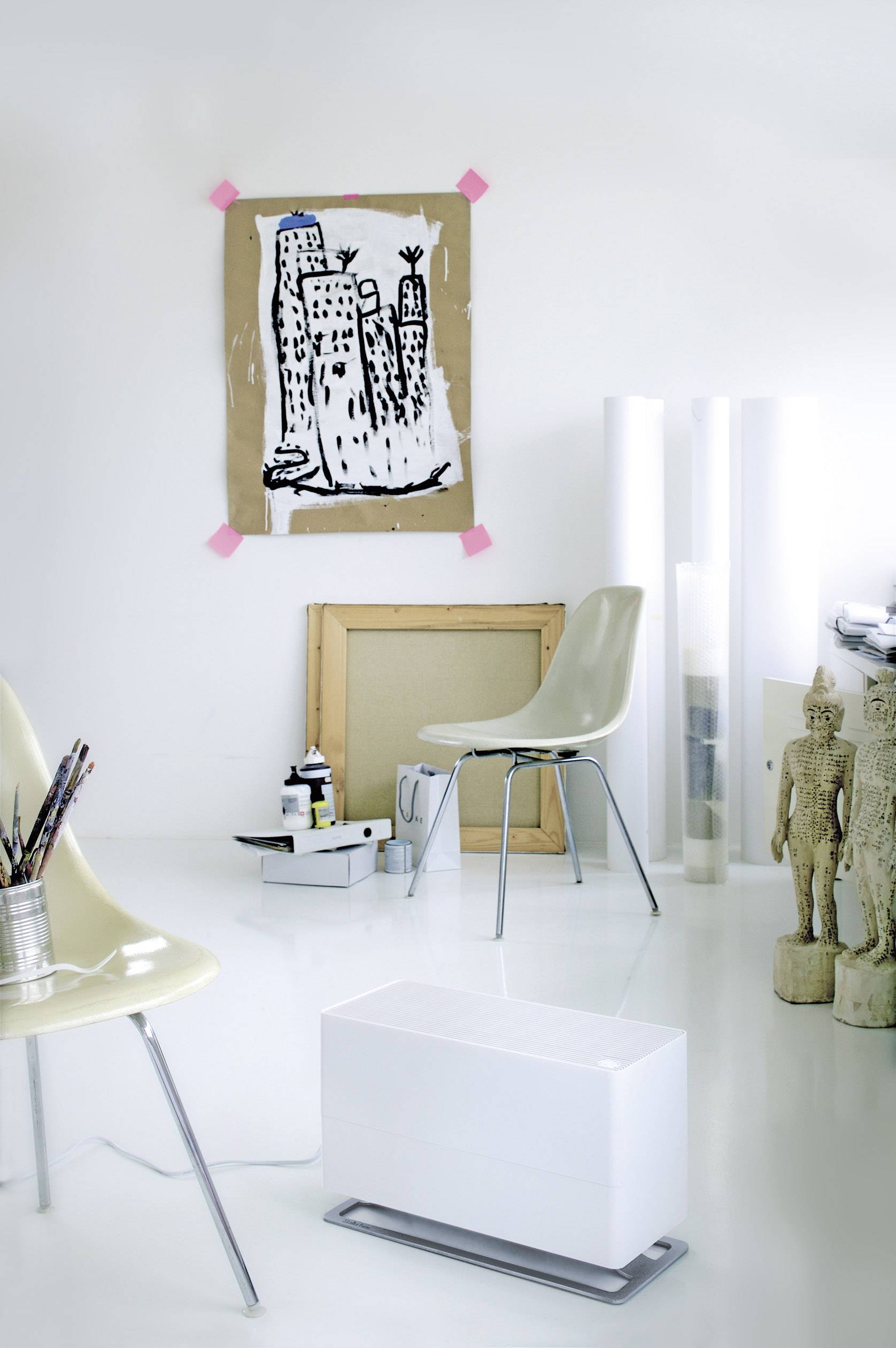 Ein heller Raum mit moderner Einrichtung. An der Wand hängt ein abstraktes Stadtbild, auf dem Boden stehen Stühle und Kunstmaterialien.