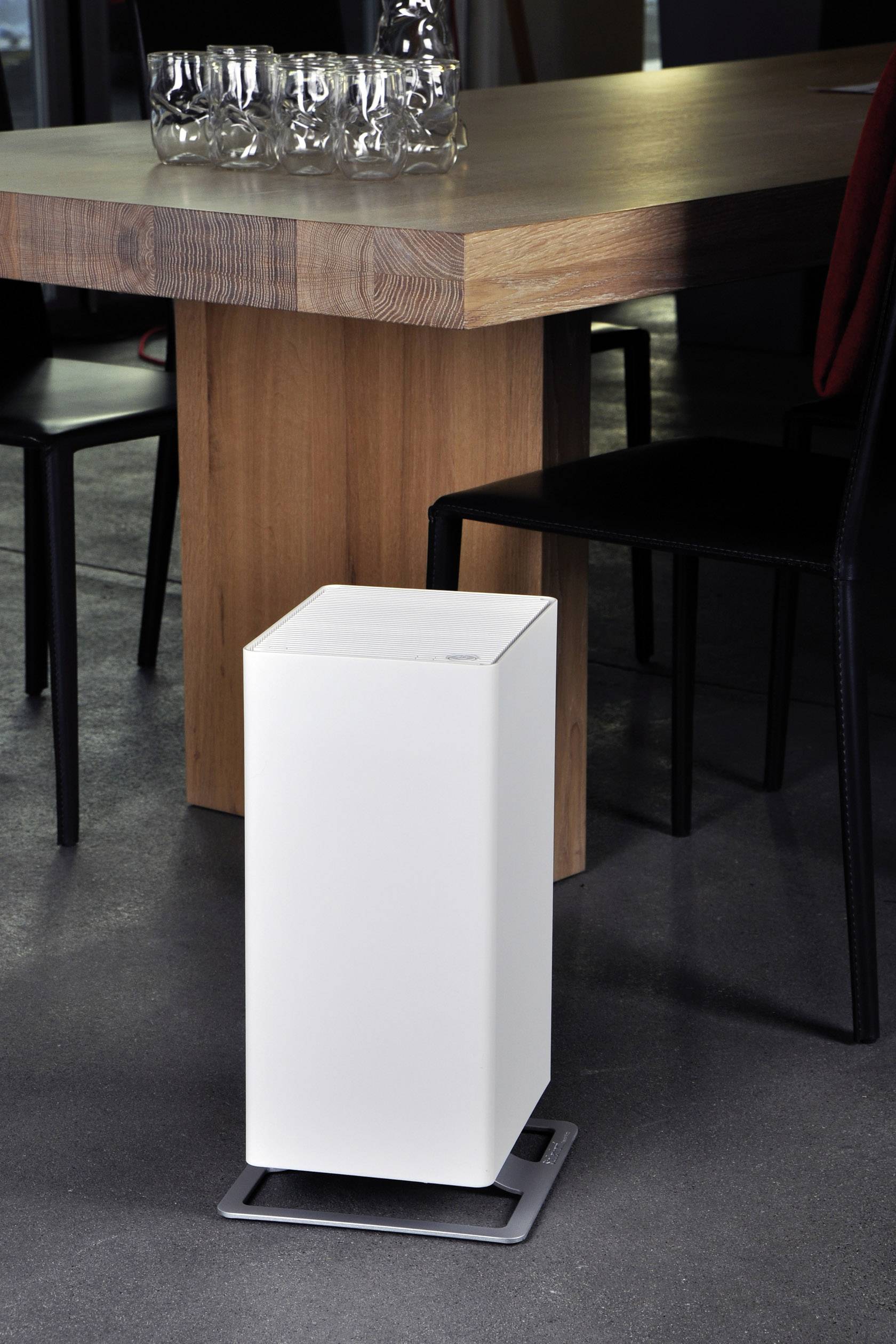 Ein weißer, rechteckiger Luftreiniger steht auf einem grauen Boden neben einem Holztisch. Darauf stehen leere Glasflaschen.