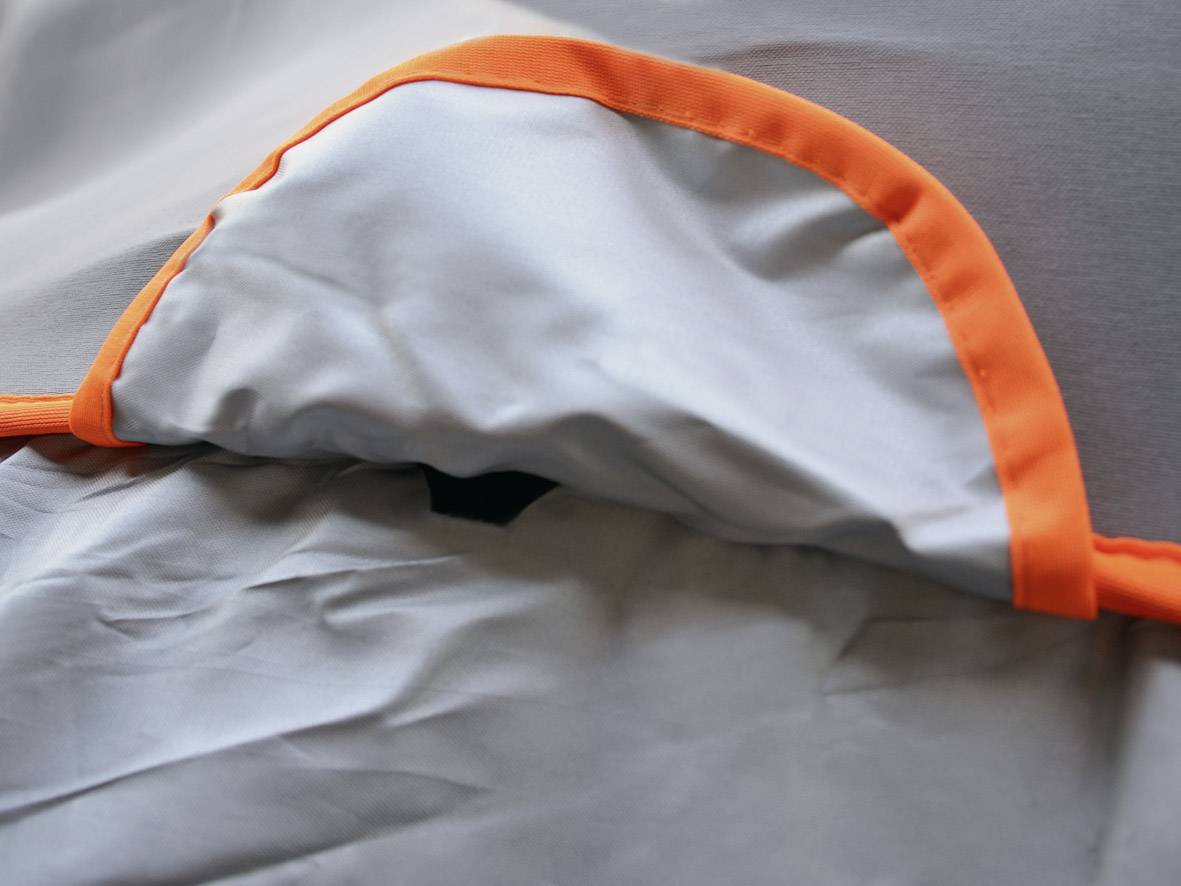 Ein graues Stoffstück mit einer leuchtend orangefarbenen Kante liegt flach. Im Hintergrund sind leichte Falten sichtbar.