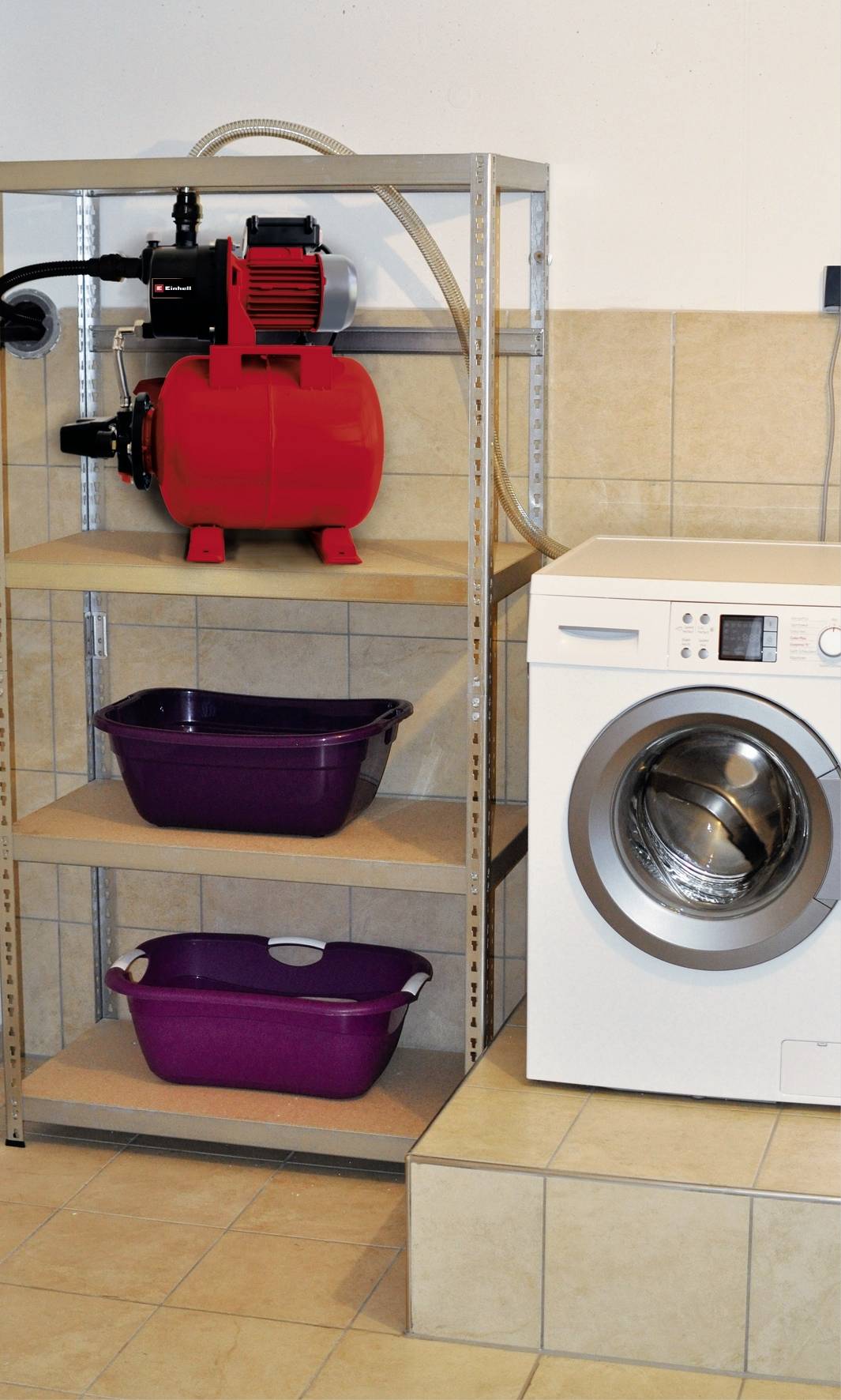 Eine weiße Waschmaschine steht auf einem gefliesten Podest. Daneben ein Regal mit einem roten Gerät oben und zwei lila Wannen unten.