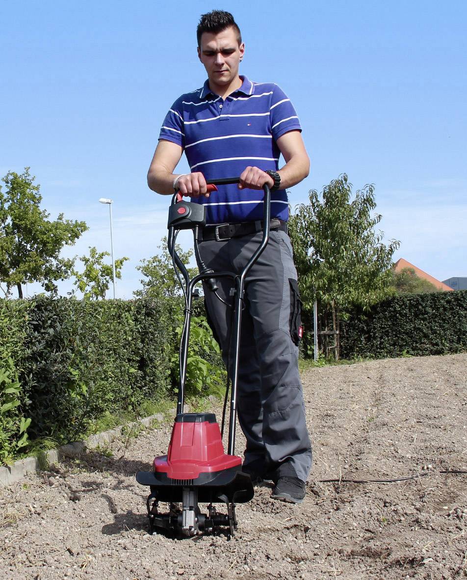 Ein Mann mit blauem gestreiftem Polohemd bedient eine rote Gartenfräse auf einem Feld. Im Hintergrund sind Bäume und ein blauer Himmel.