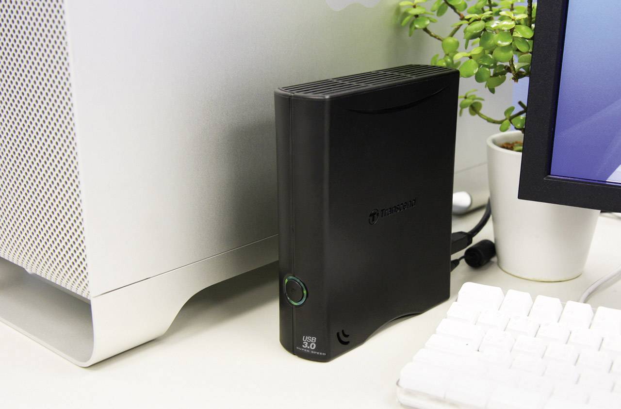 Externe Festplatte neben einem Computer, mit Pflanzen in der Nähe; dient zur Datenlagerung und Backup-Erweiterung.