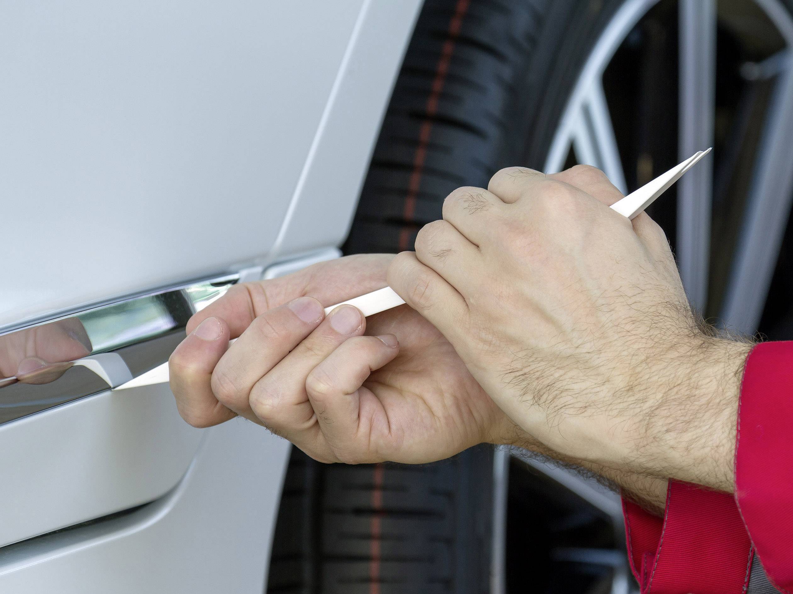 Detailarbeit an einem weißen Auto. Eine Person benutzt ein Kunststoffwerkzeug, um Teile der Karosserie oder Zierleiste zu bearbeiten.