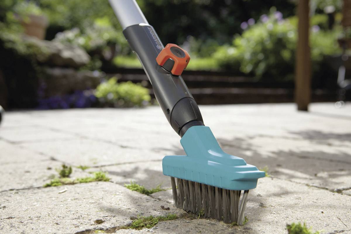 Ein Bürstengerät mit langem Griff entfernt Unkraut aus Ritzen in einer Terrasse aus Steinplatten, umgeben von Gartenpflanzen.