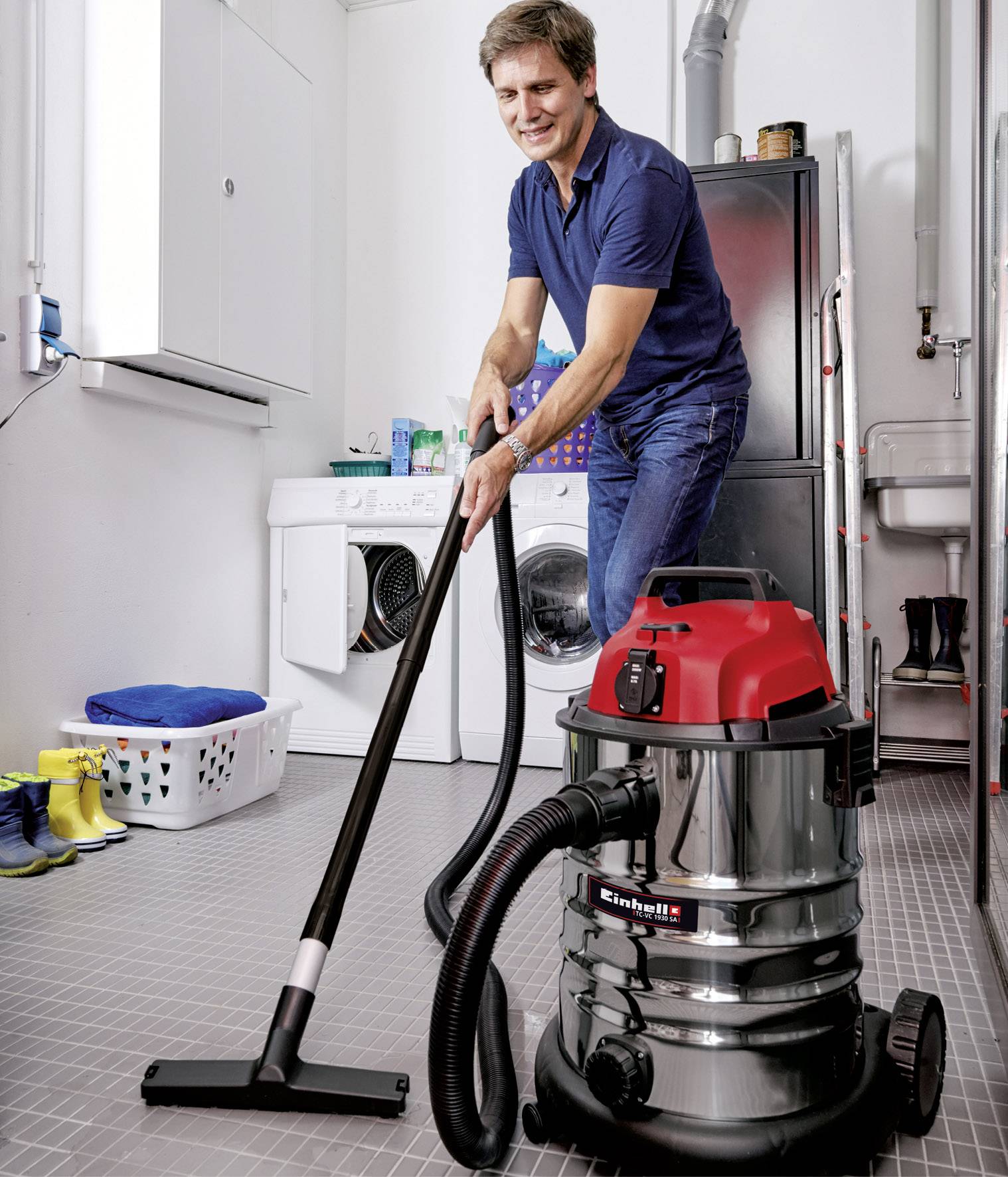 Ein Mann in einem blauen Hemd saugt mit einem Industriesauger einen grauen Boden in einem Hauswirtschaftsraum. Im Hintergrund sind Waschmaschine und Stiefel.