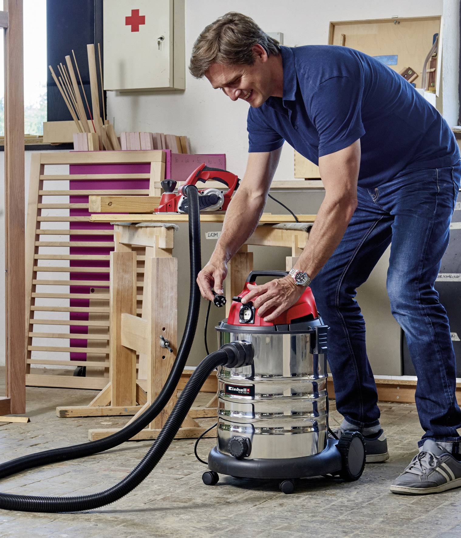 Mann in Werkstatt bedient einen Industriesauger mit rotem Deckel, umgeben von Holzmaterialien und Werkzeugen.