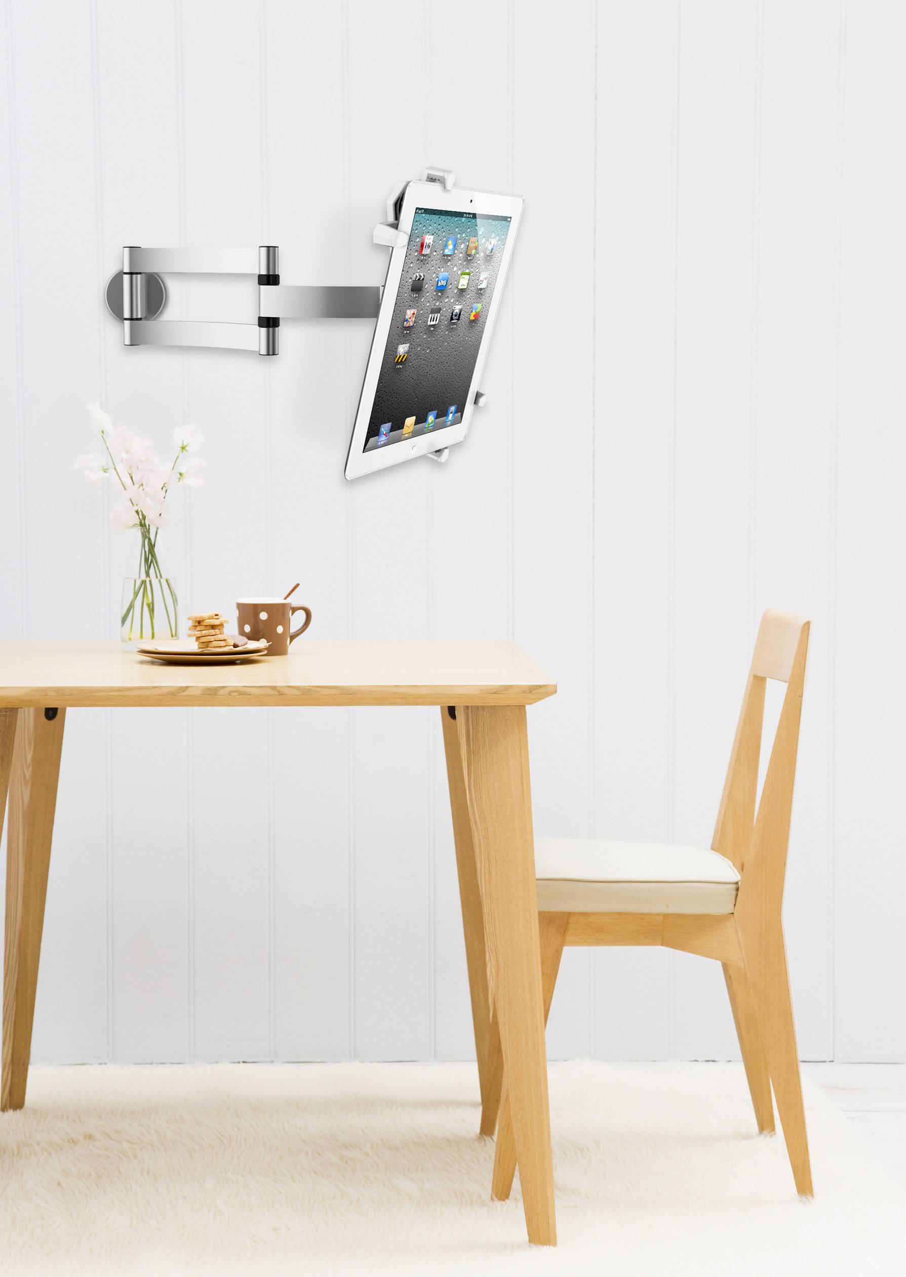 Tablet an einer beweglichen Wandhalterung über einem Holztisch mit Vase und Tasse, daneben ein Holzstuhl vor heller Wand.