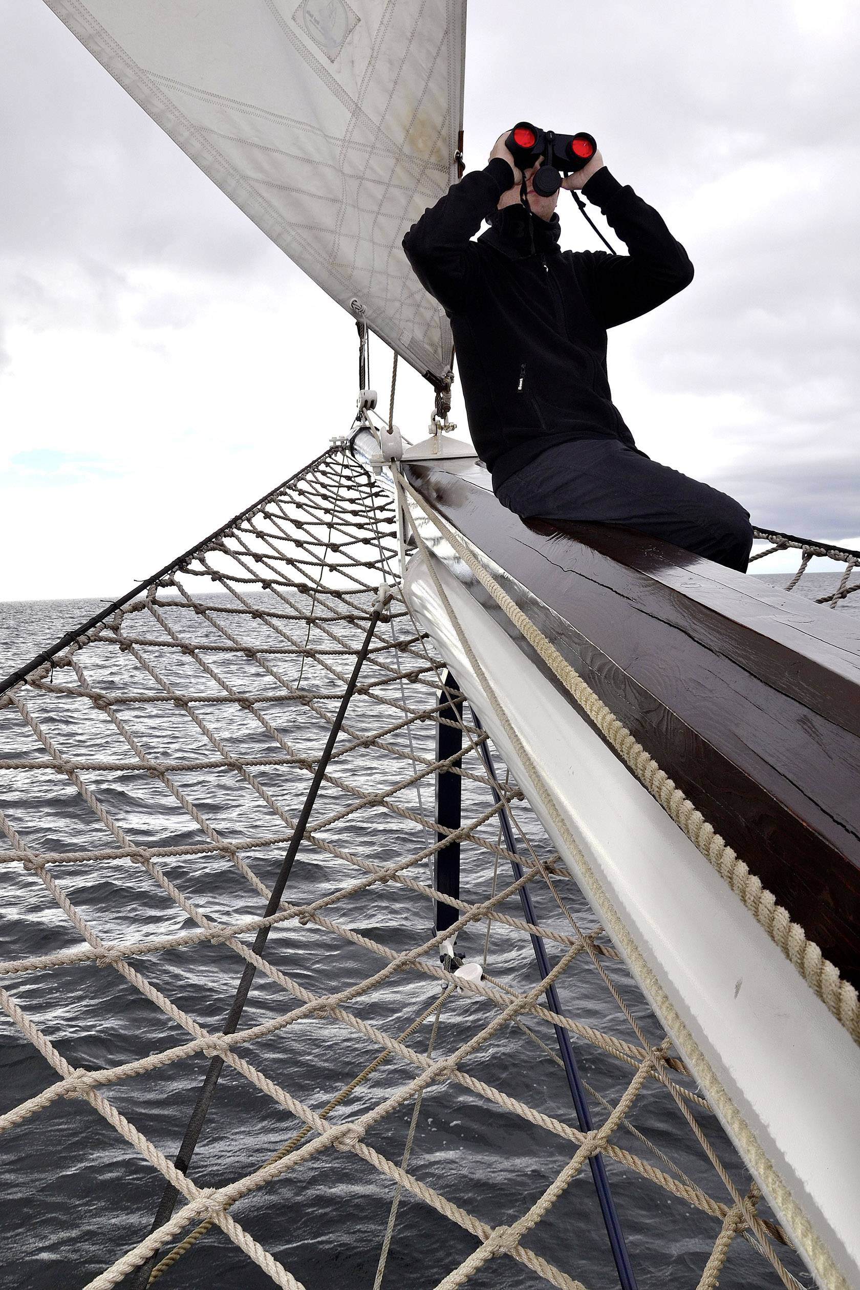 Eine Person sitzt auf einem Segelbootbug und schaut mit Fernglas aufs Meer. Der Himmel ist bewölkt, und das Meer ist ruhig.