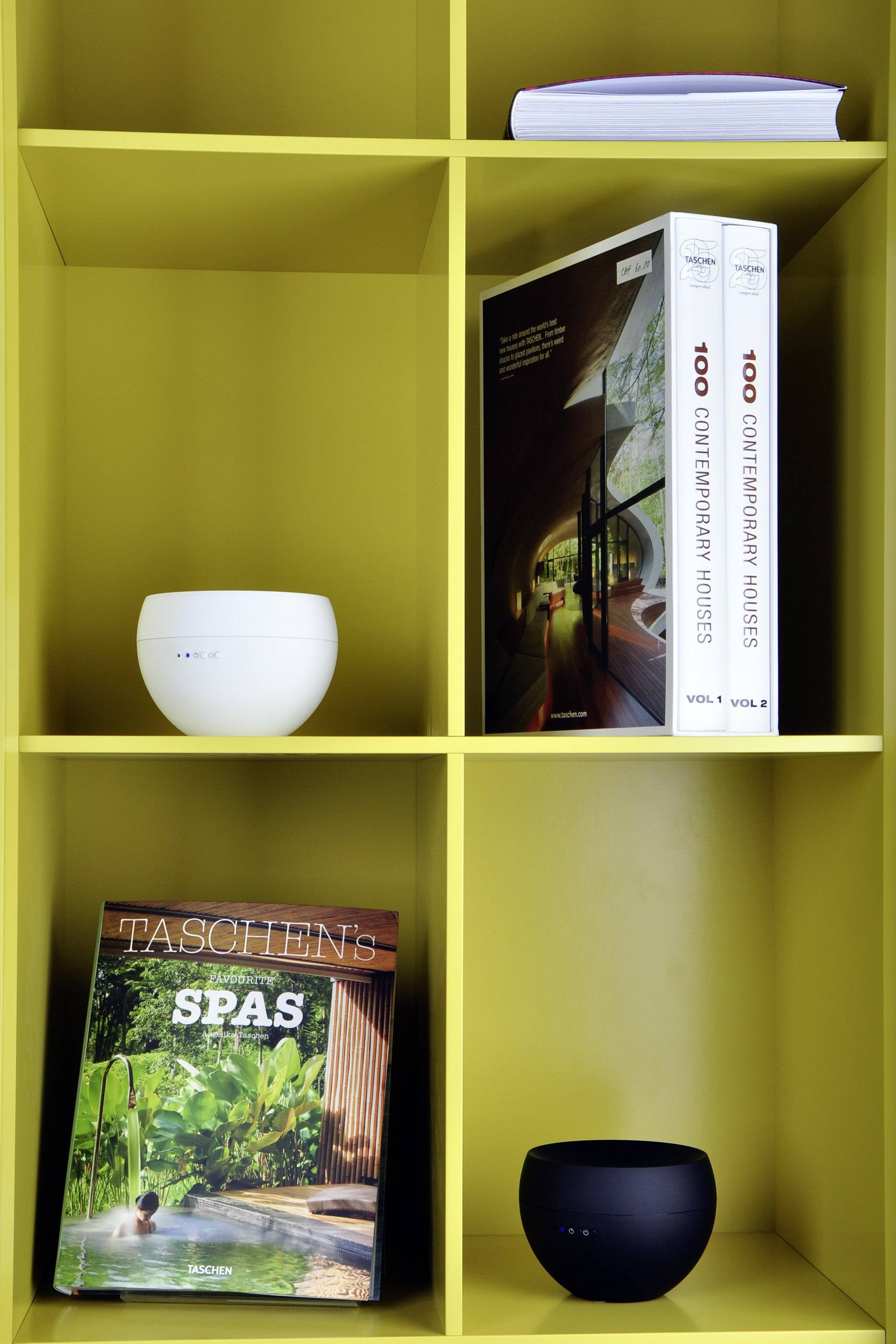 Ein gelbes Regal mit verschiedenen Fächern, in denen Bücher, ein weißer und ein schwarzer Lautsprecher sowie ein Katalog platziert sind.