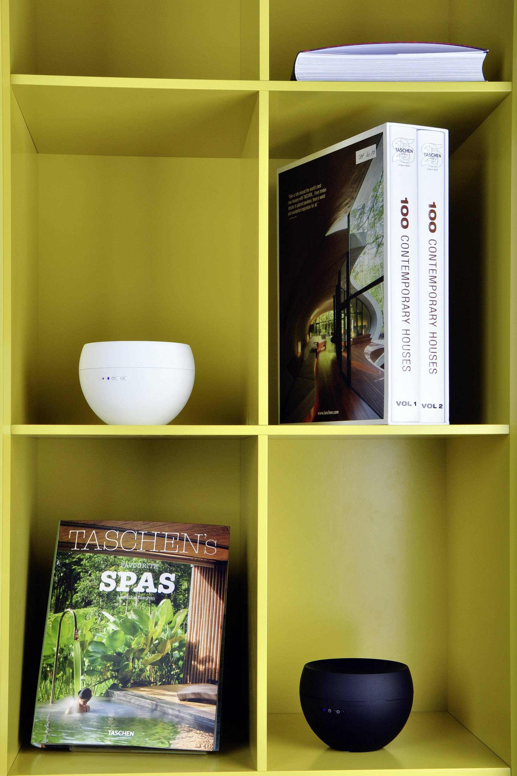 Ein gelbes Regal mit zwei Büchern '100 Contemporary Houses', einer Zeitschrift 'TASCHEN's SPAS' und zwei runden Lautsprechern.