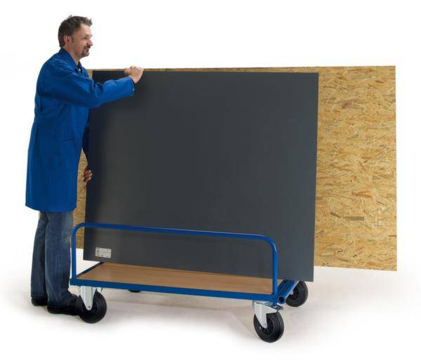 Ein Mann in einem blauen Mantel transportiert eine große graue und holzfarbene Platte auf einem blauen Rollwagen.