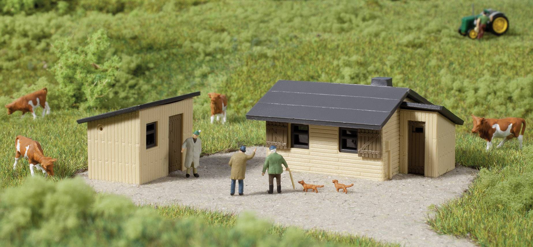 Kleines Modellbauernhof-Diorama mit zwei Gebäuden, Figuren, zwei Hunden und Kühen auf grünem Feld. Ein Traktor ist im Hintergrund.