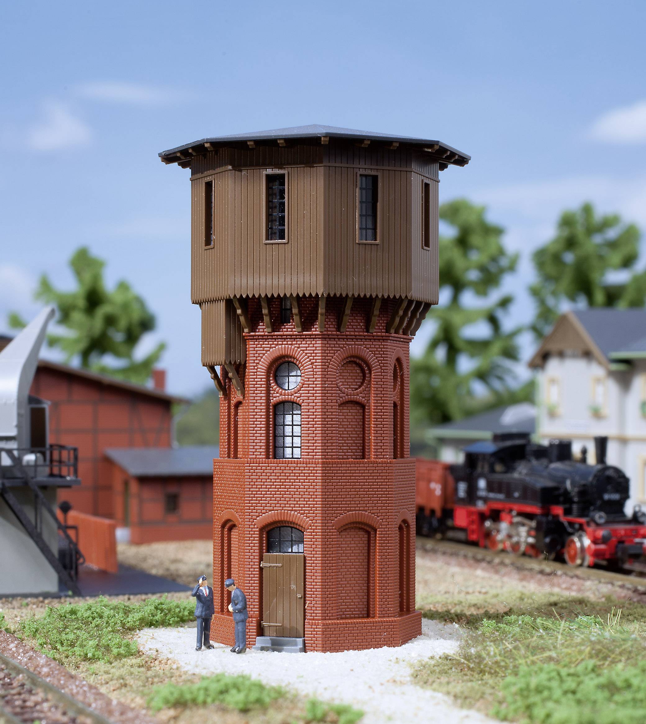 Ein Modell eines Wasserturms aus rotem Backstein mit hölzernem Obergeschoss, umgeben von Eisenbahnmodelllandschaft, zwei Figuren davor.