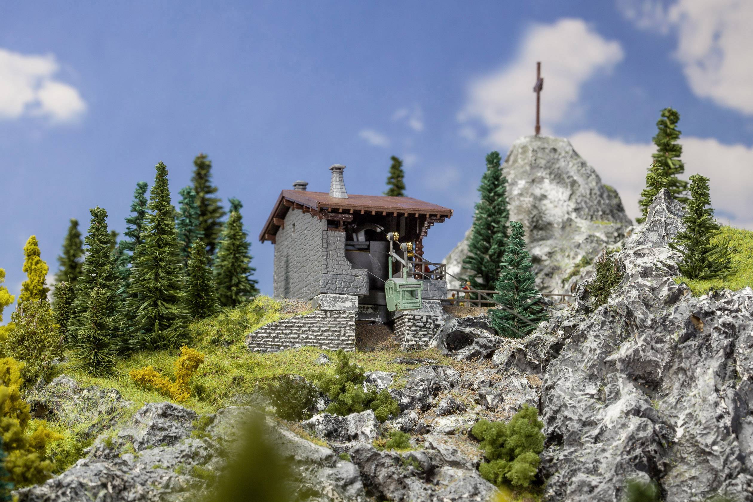 Eine Berghütte in alpiner Landschaft mit umliegenden Bäumen und Felsen, ein Gipfelkreuz steht auf einem Berg im Hintergrund unter blauem Himmel.