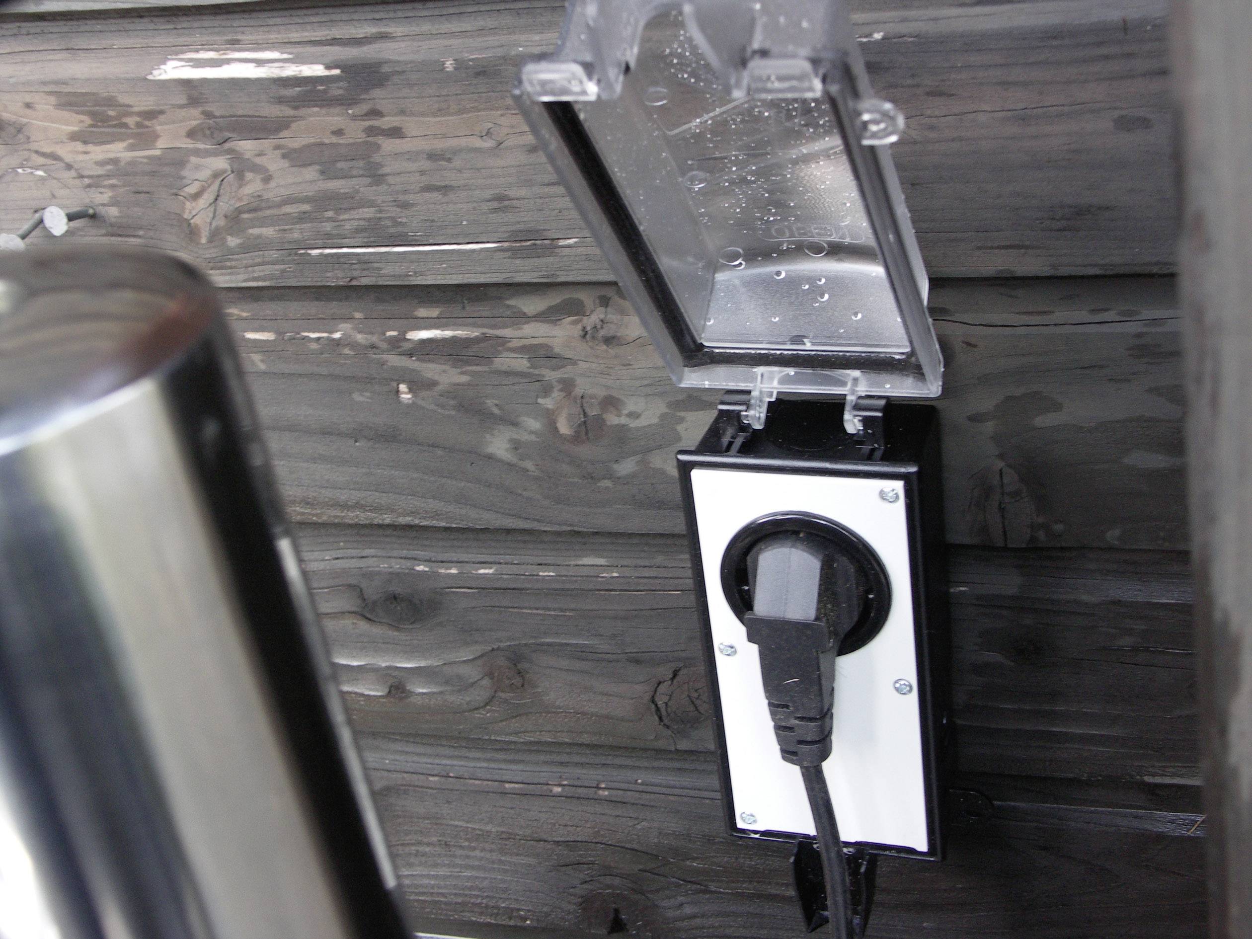 Eine Außensteckdose ist an einer Holzoberfläche montiert. Der Schutzdeckel ist geöffnet und ein schwarzes Kabel ist eingesteckt.