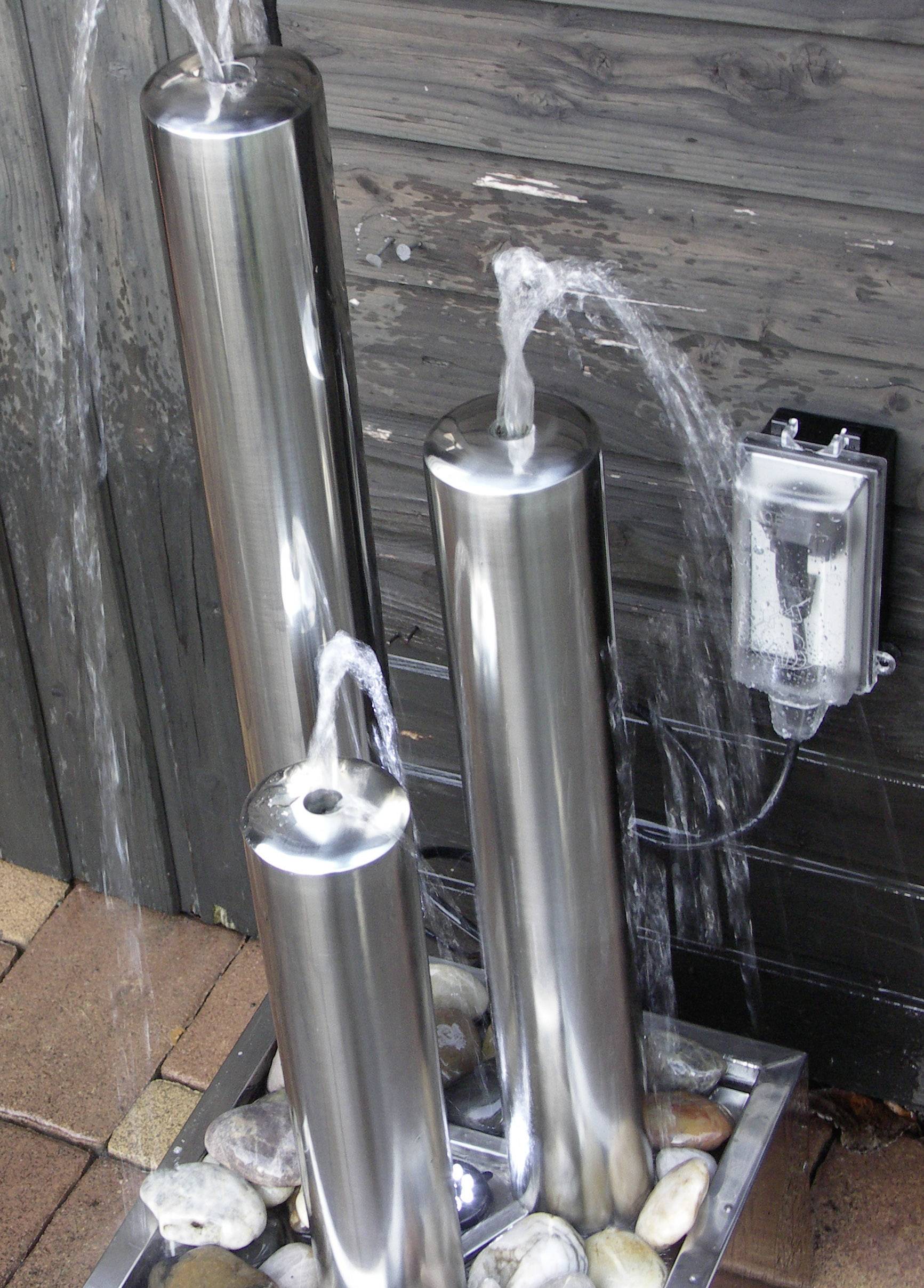 Drei silberne Wasserfontänen unterschiedlicher Höhe sprühen Wasser in einem Gartenbereich, umgeben von Steinen.