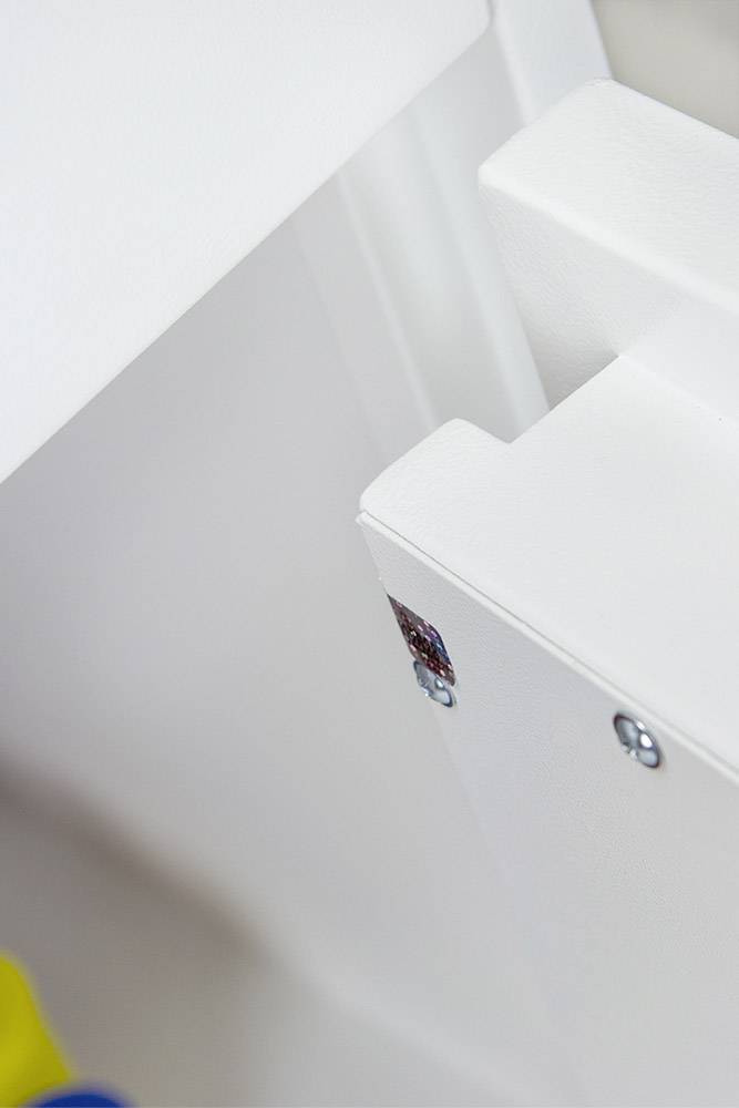 Nahaufnahme eines weißen Möbelstücks mit einem Metallwinkel in der Ecke und sichtbaren Schrauben. Links unten ist ein verschwommenes gelbes Element.