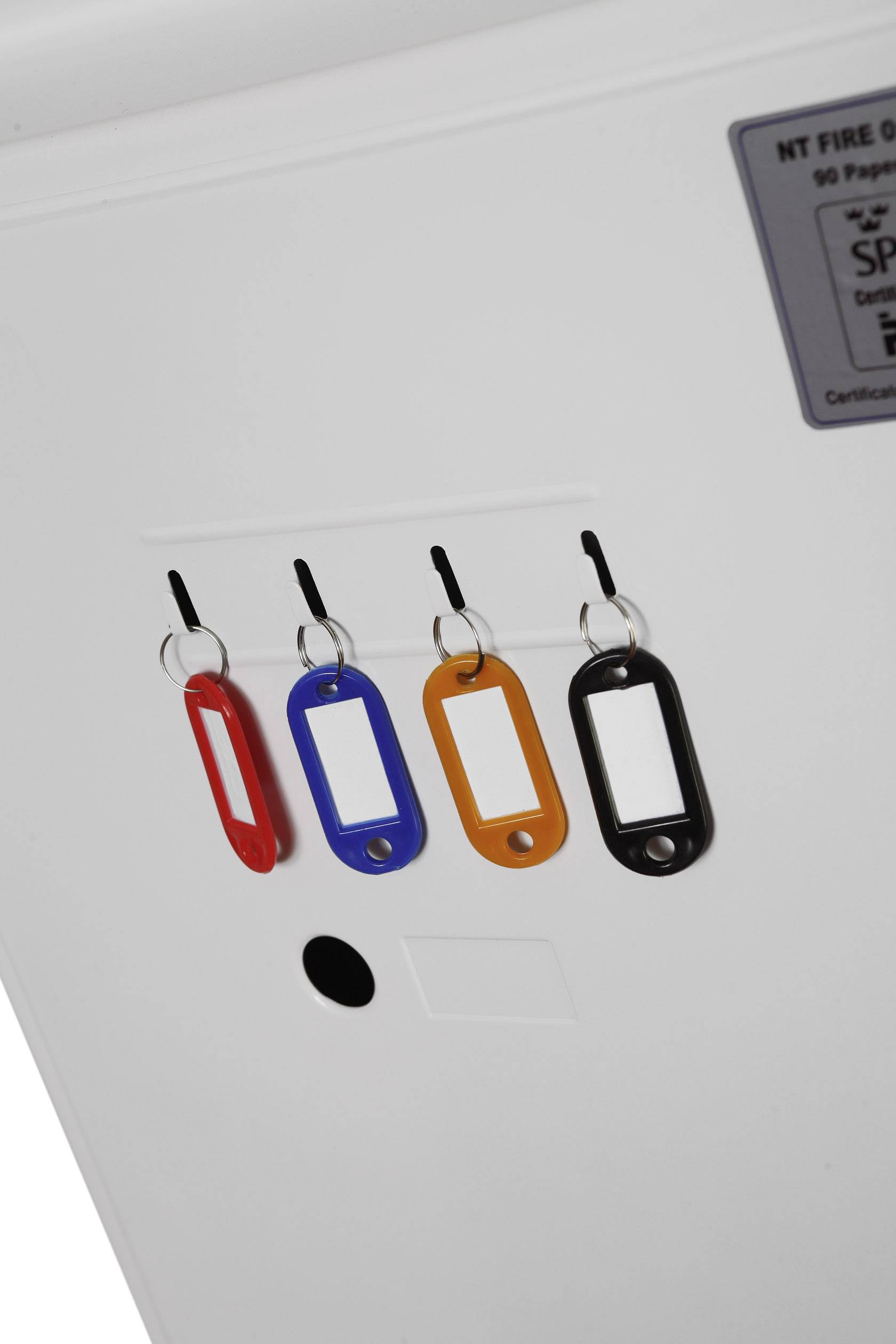 Vier farbige Schlüsselanhänger (rot, blau, orange, schwarz) hängen an Haken an einer weißen Oberfläche.