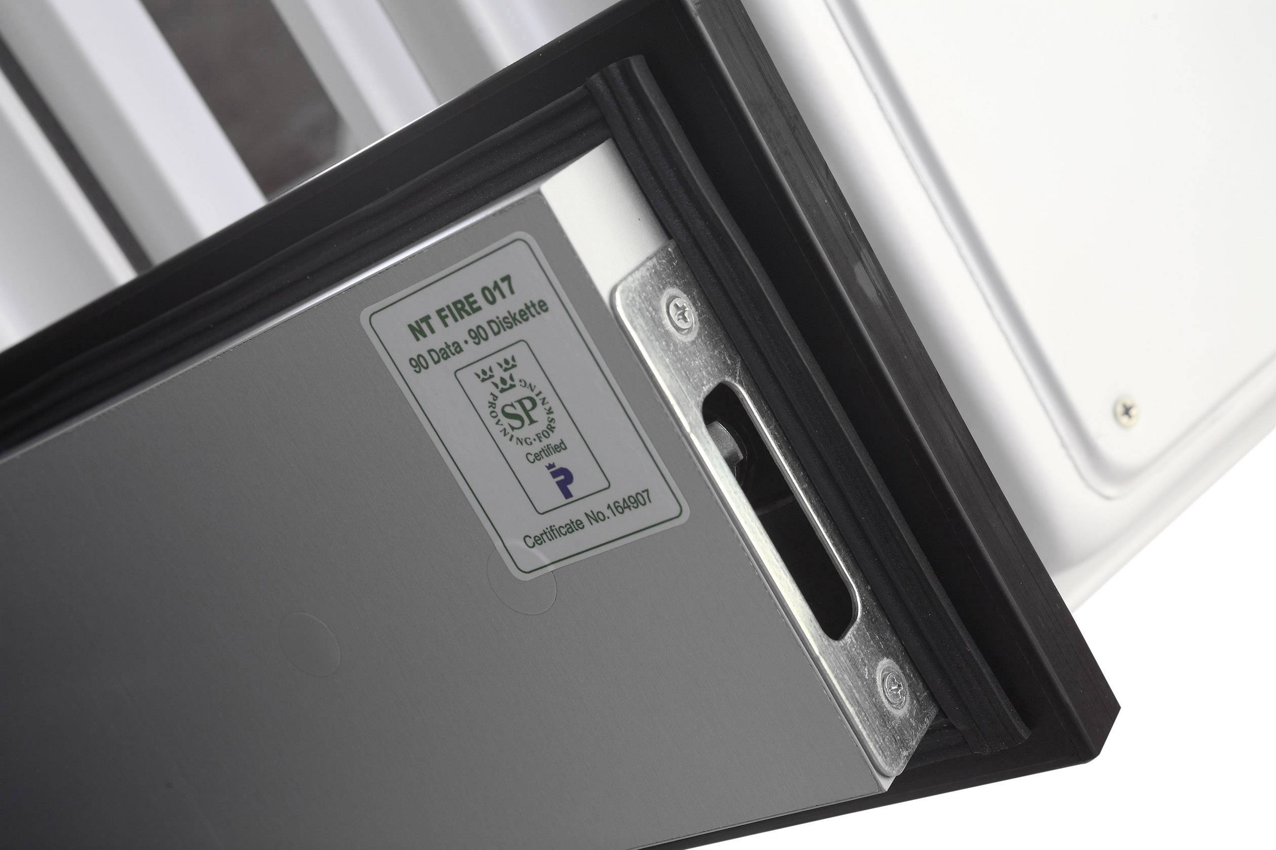 Nahaufnahme eines Metallschlosses mit Zertifikatsetikett auf der Innenseite einer grauen Tür. Links sind Scharniere sichtbar.