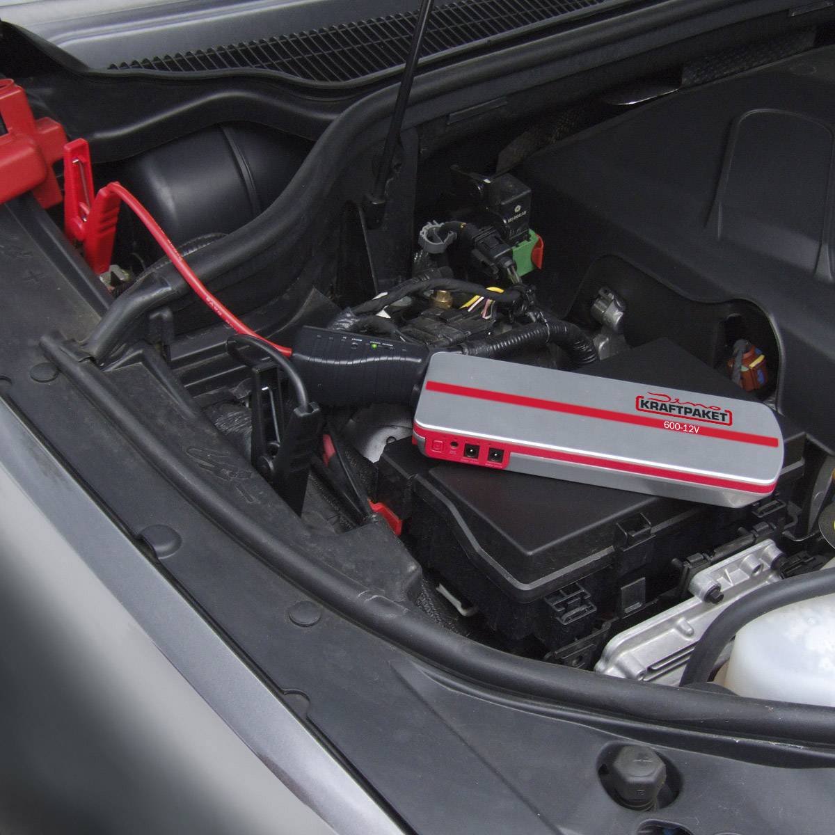 Auto-Motorraum mit einem Starthilfegerät, das an die Batterie angeschlossen ist. Das Gerät liegt sicher auf der Batterieabdeckung.