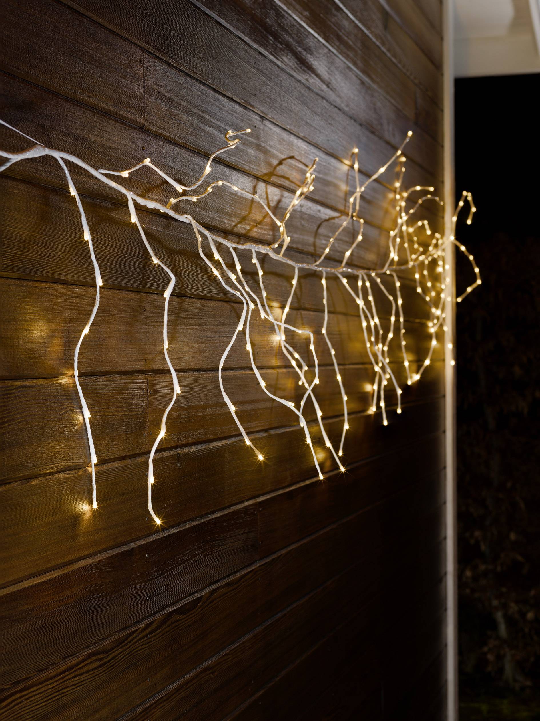 Lichterkette in Form von Zweigen dekoriert eine Holzfassade im Dunkeln. Die warmweißen Lichter schaffen eine gemütliche Atmosphäre.