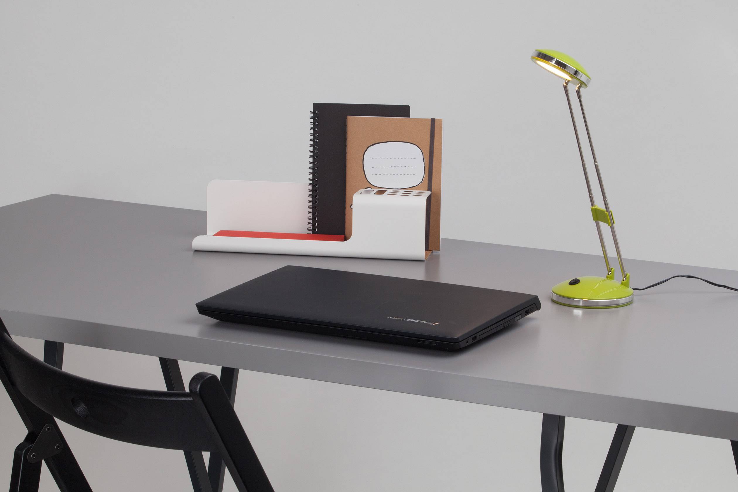 Ein grauer Schreibtisch mit einem schwarzen Laptop, einer Tischlampe und einer Ablage mit Heften und Notizbüchern.