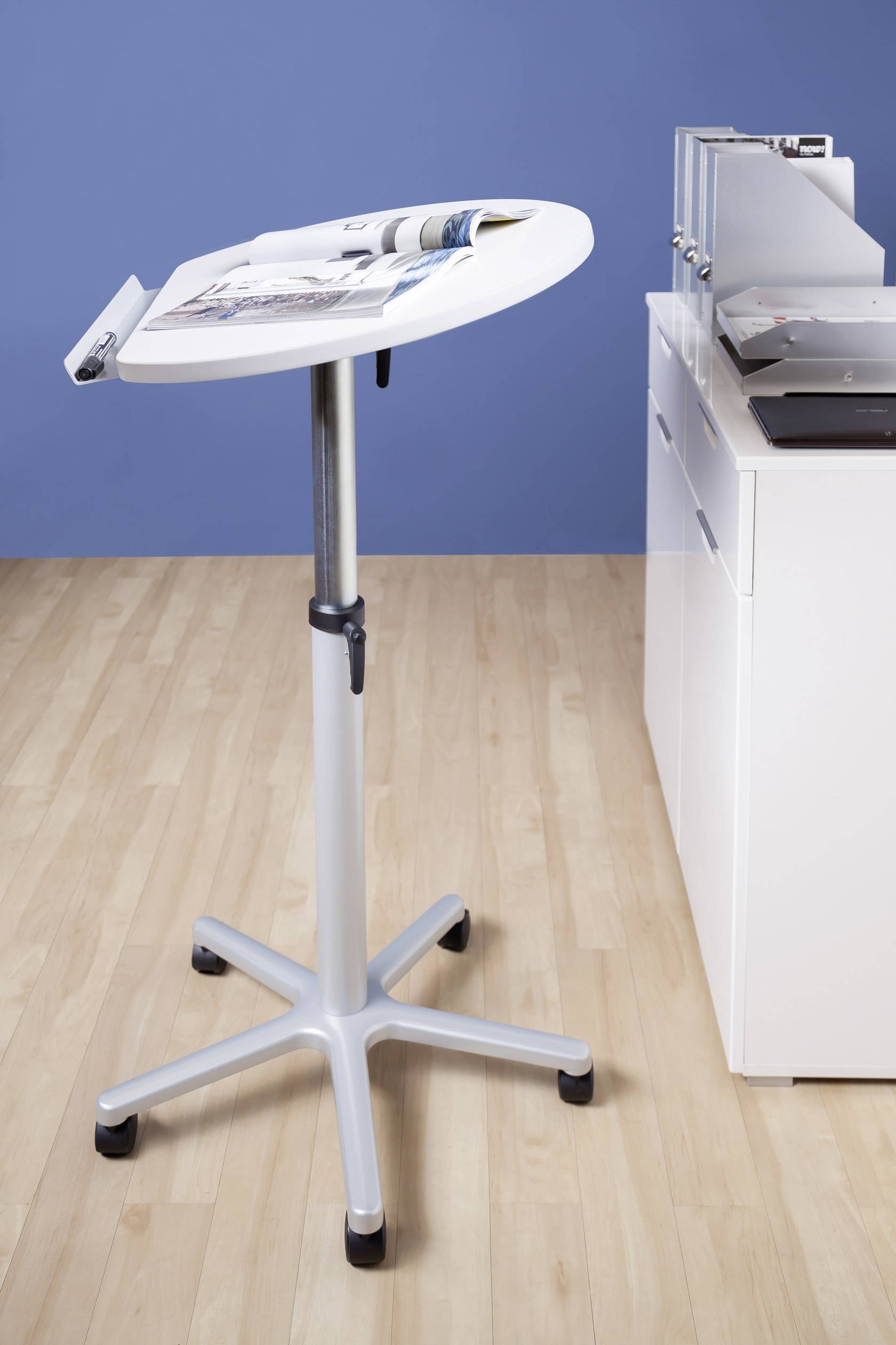 Ein höhenverstellbarer Beistelltisch aus Metall mit Rollen, beladen mit Zeitschriften, steht neben einem weißen Aktenschrank auf einem Holzfußboden.