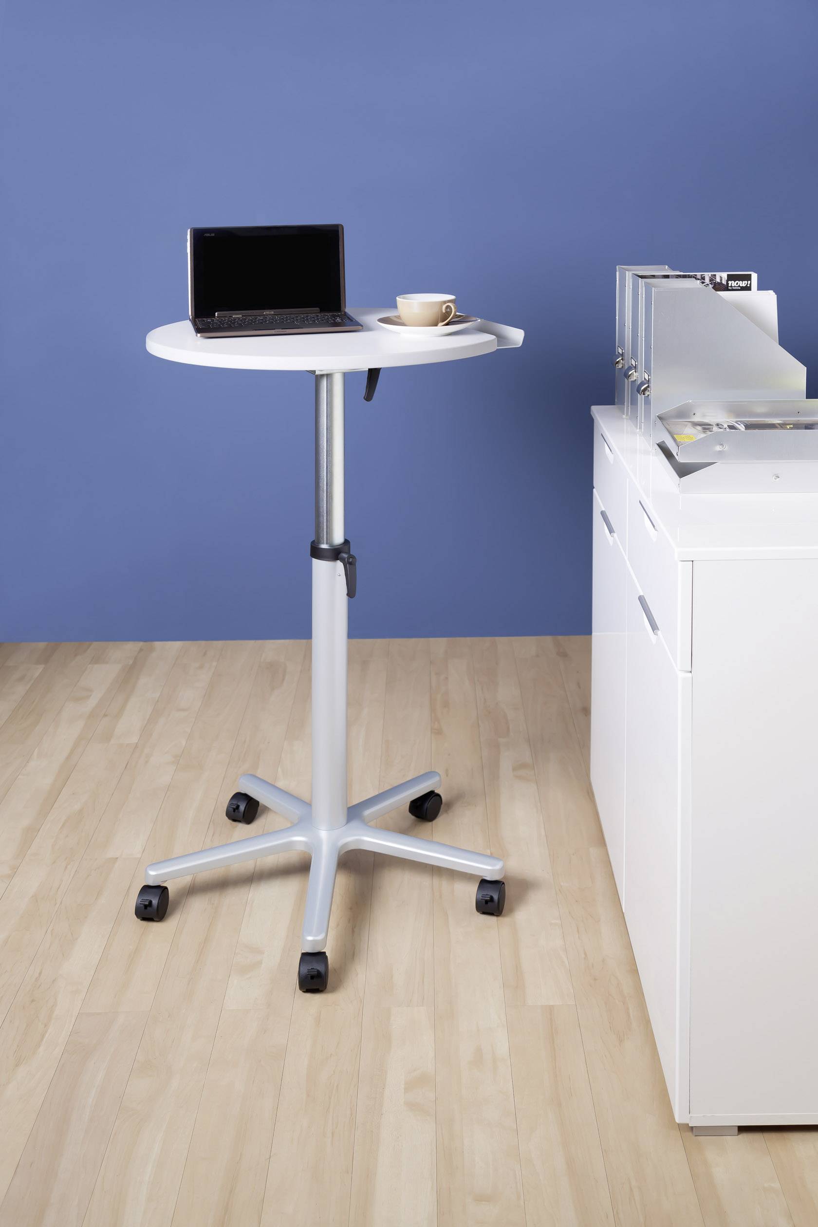 Runder, höhenverstellbarer Stehtisch auf Rollen mit Laptop und Kaffeetasse, daneben ein weißes Schrankmodul vor einer blauen Wand.